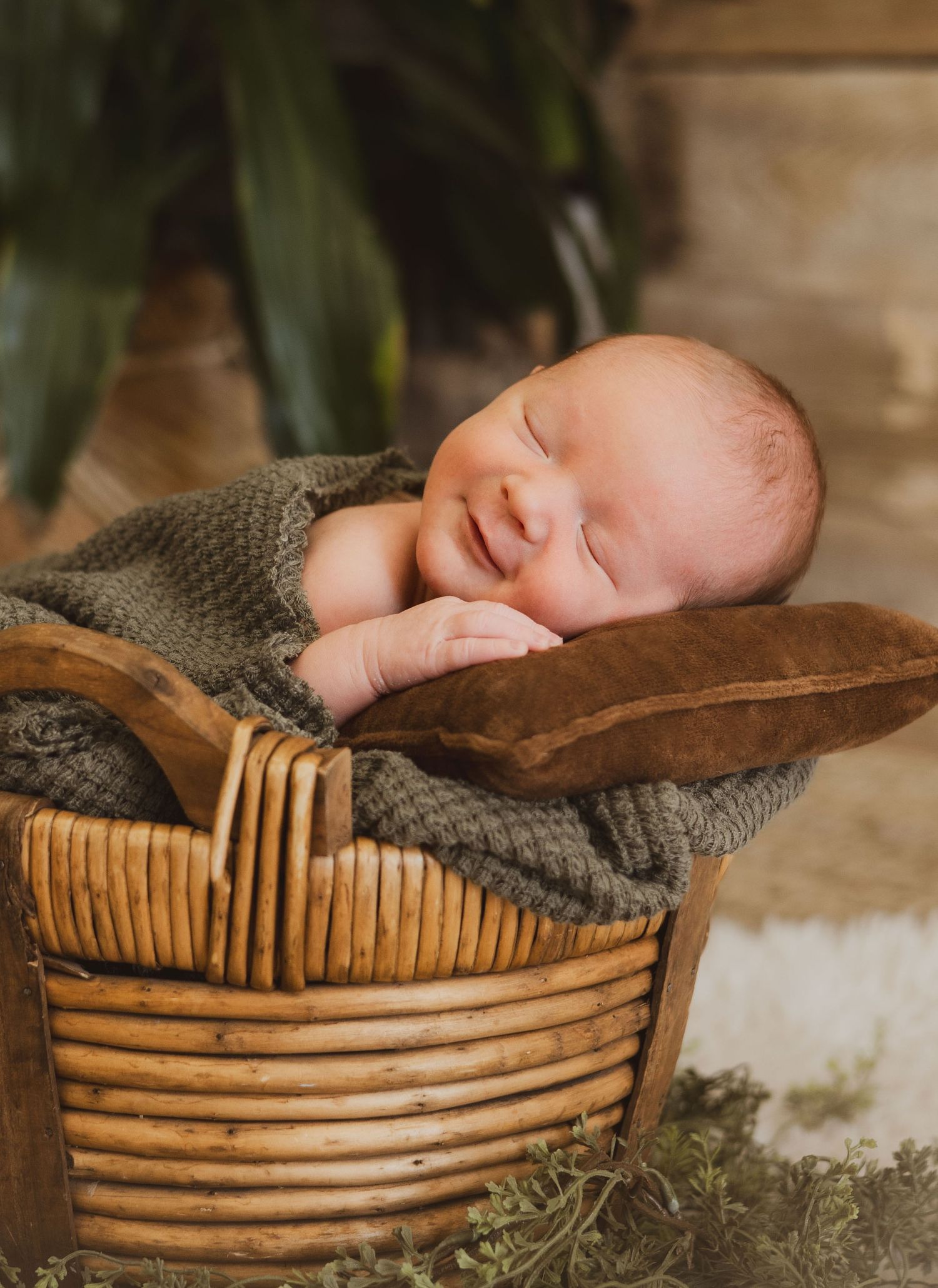 Smiling newborn baby sleeping in a rustic wooden basket with green props.