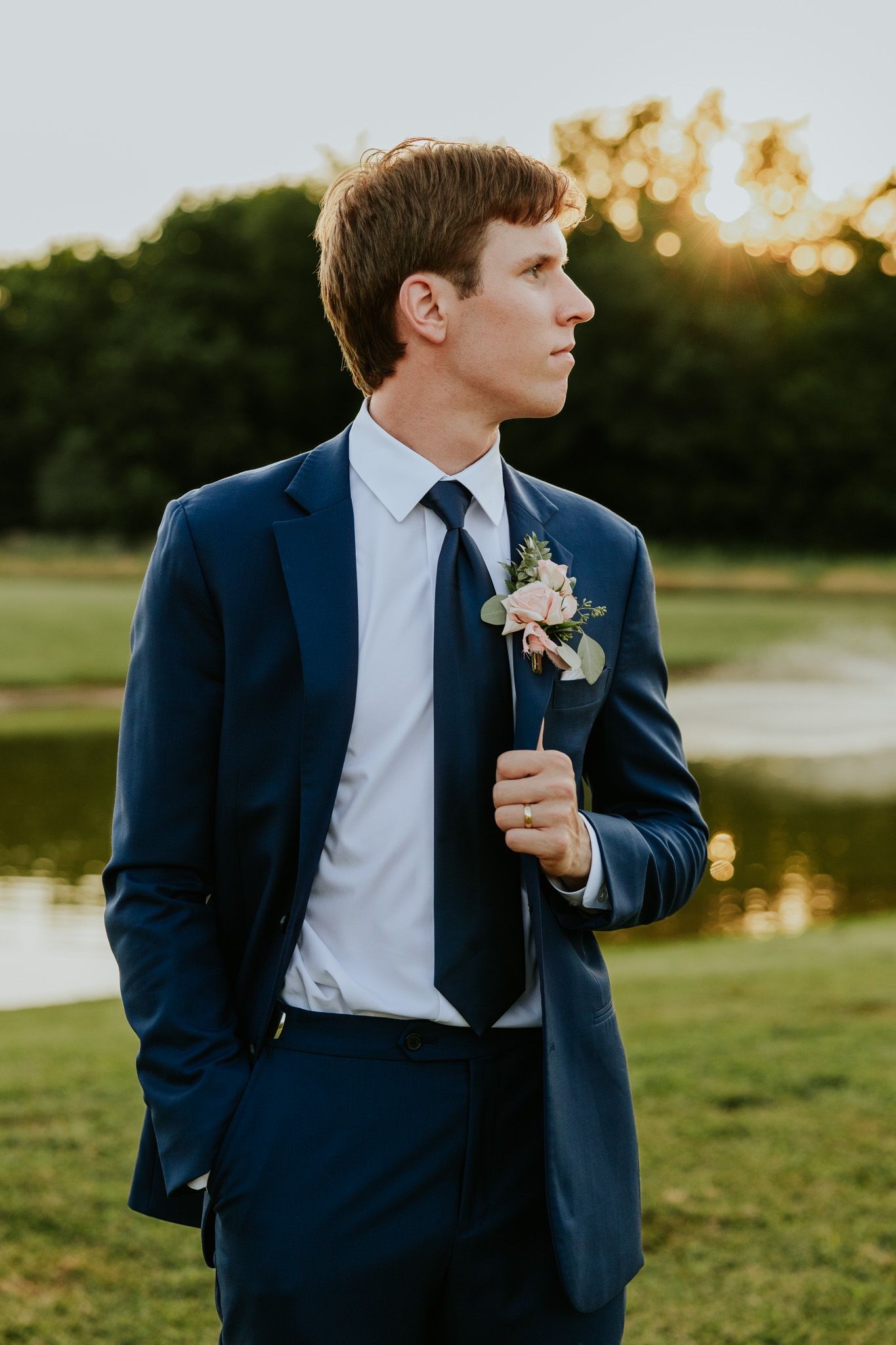 Groom in navy suit with floral boutonniere posing outdoors at sunset near a pond.
