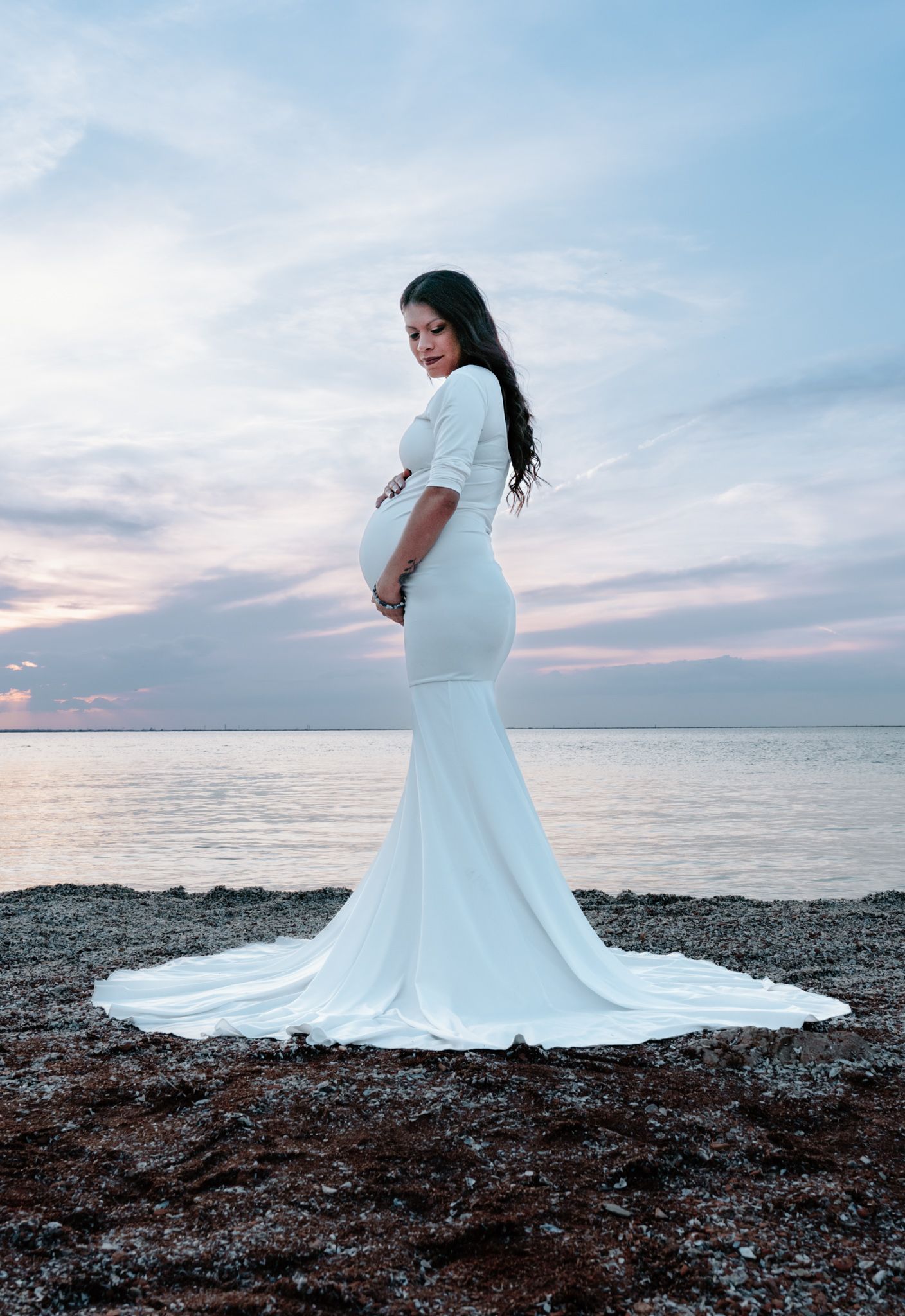 Pregnant woman in elegant white mermaid gown standing on rocky beach at sunset over calm water.