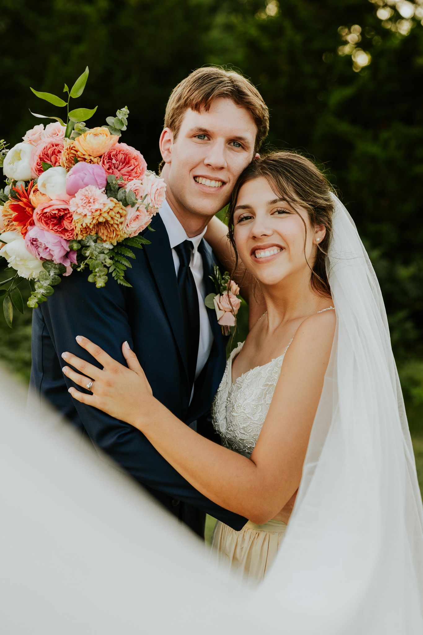 A smiling bride and groom pose together outdoors, she holds a colorful pink and peach bouquet, wearing a white gown and veil.