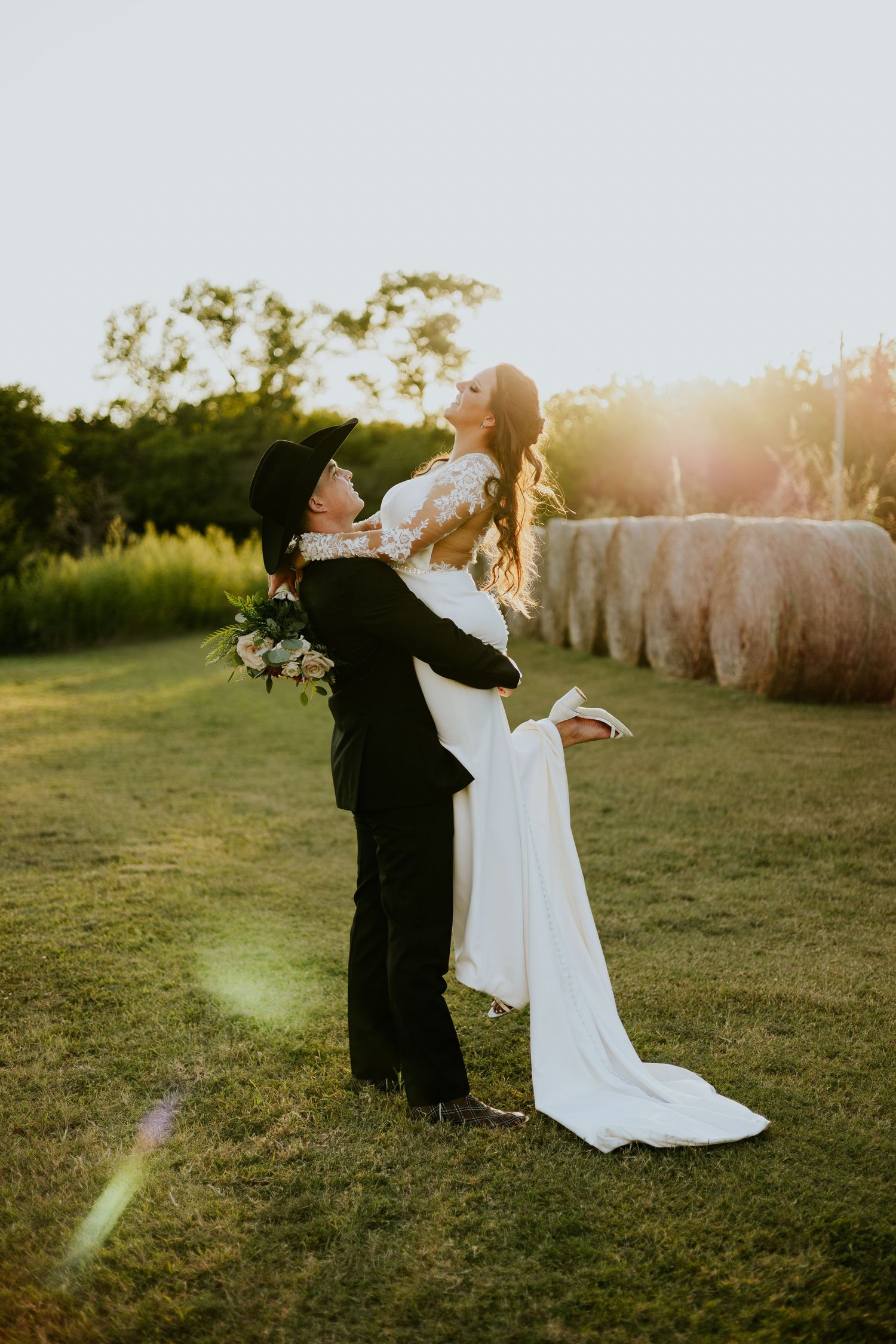 Groom in black suit lifting bride in white dress at sunset near hay bales.