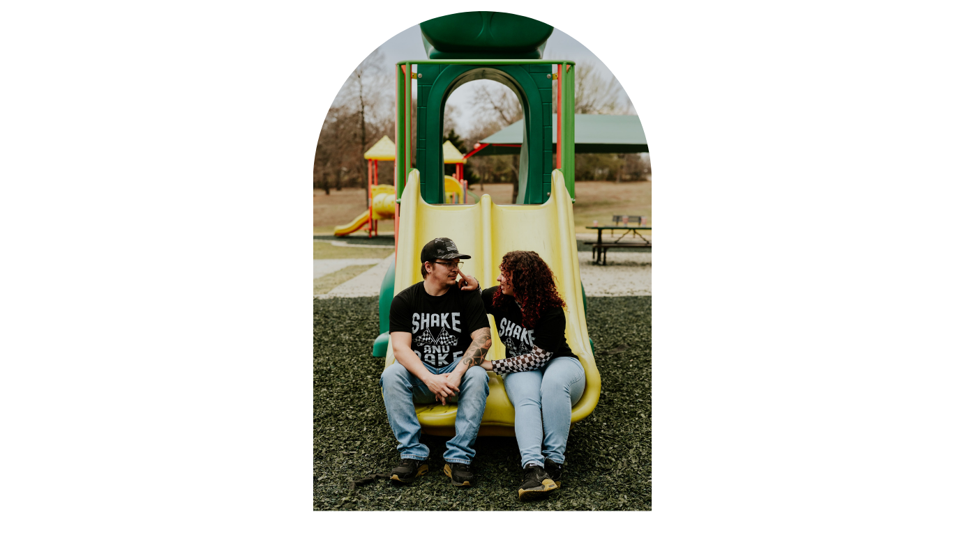 Couple on yellow playground slide smiling at each other, framed in arch-shaped photo edit.