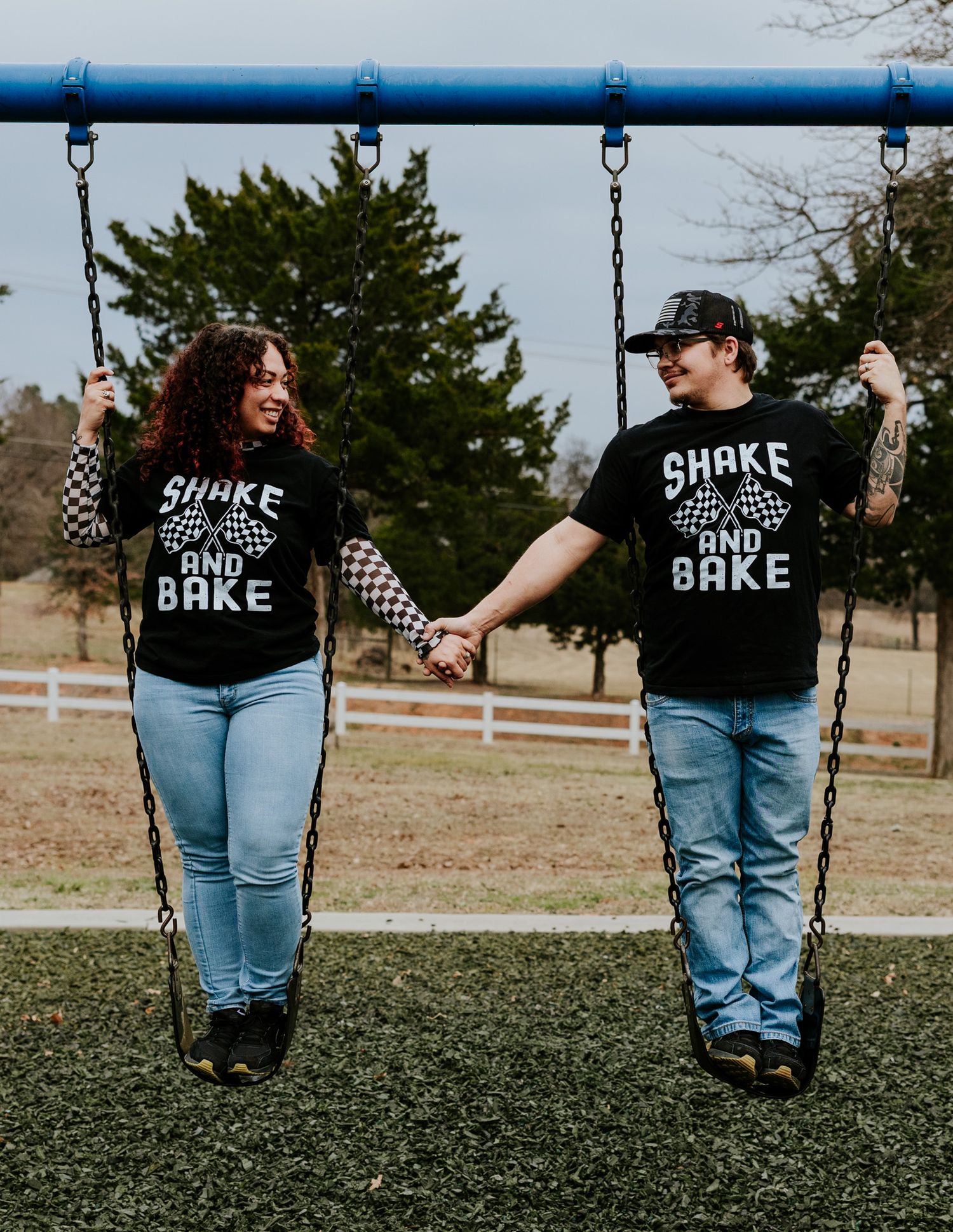 Couple holding hands on swings wearing matching Shake and Bake black t-shirts and jeans in a park.