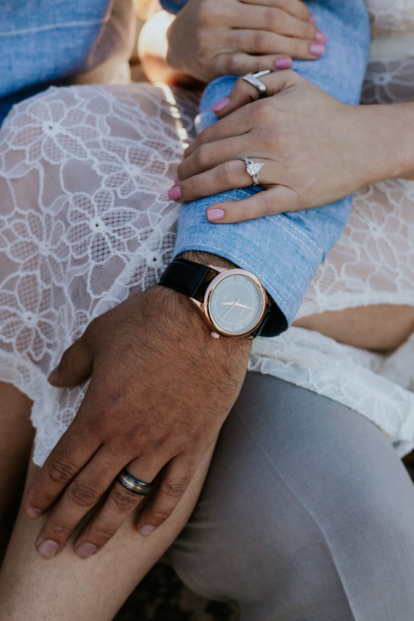 Close-up of couple's hands showing diamond engagement ring, wedding band, and elegant rose gold watch.