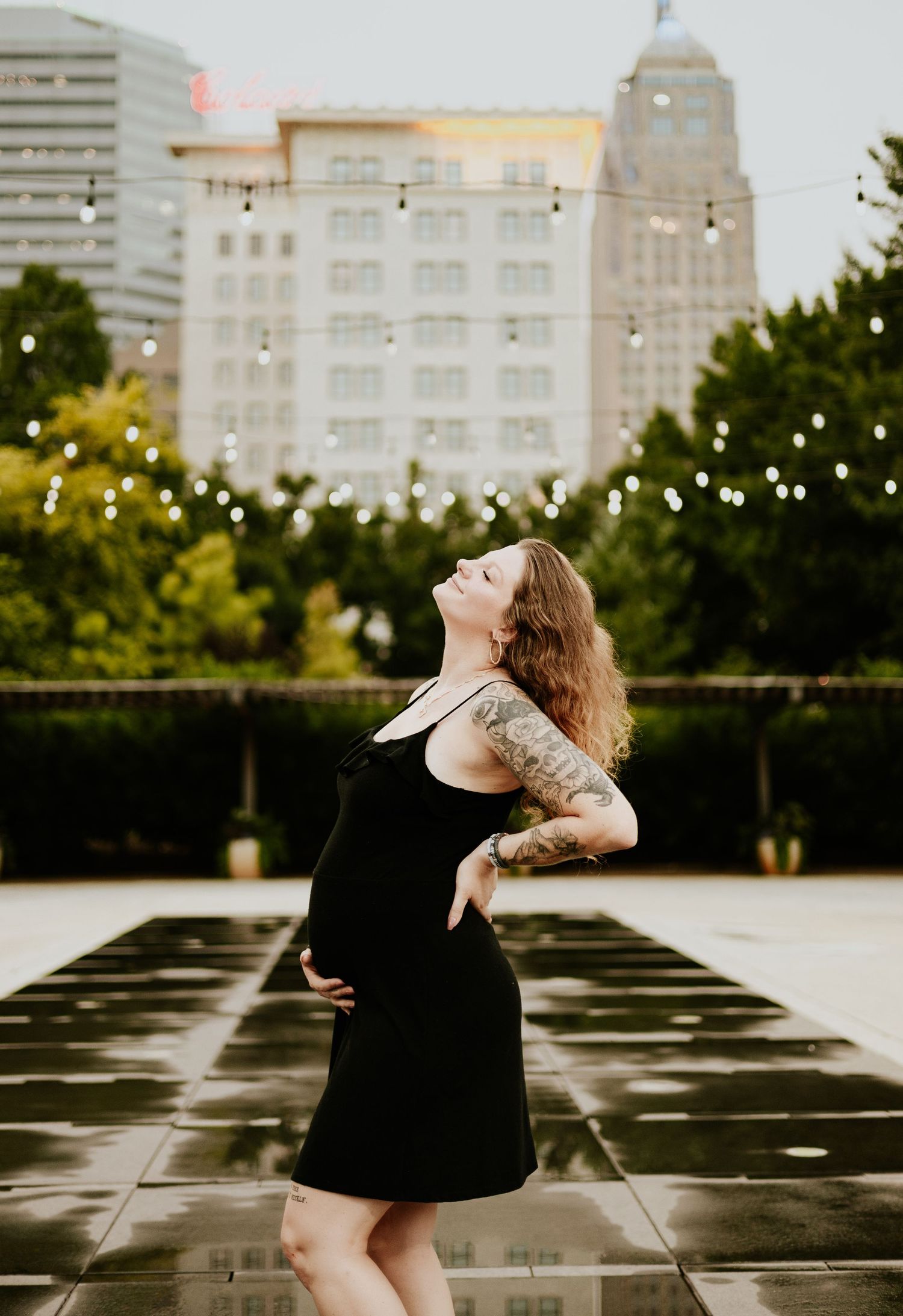 A pregnant tattooed woman in a black dress poses gracefully on a rooftop with a city skyline backdrop.