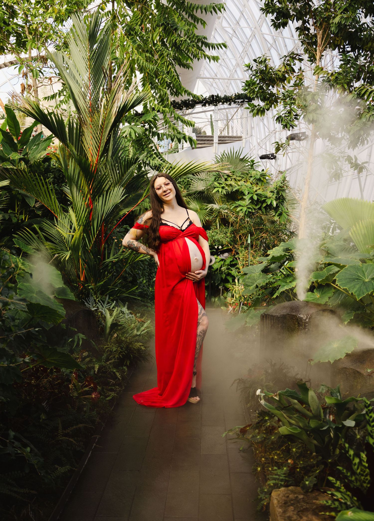 Radiant pregnant woman in a flowing red gown posing in a misty tropical greenhouse setting.