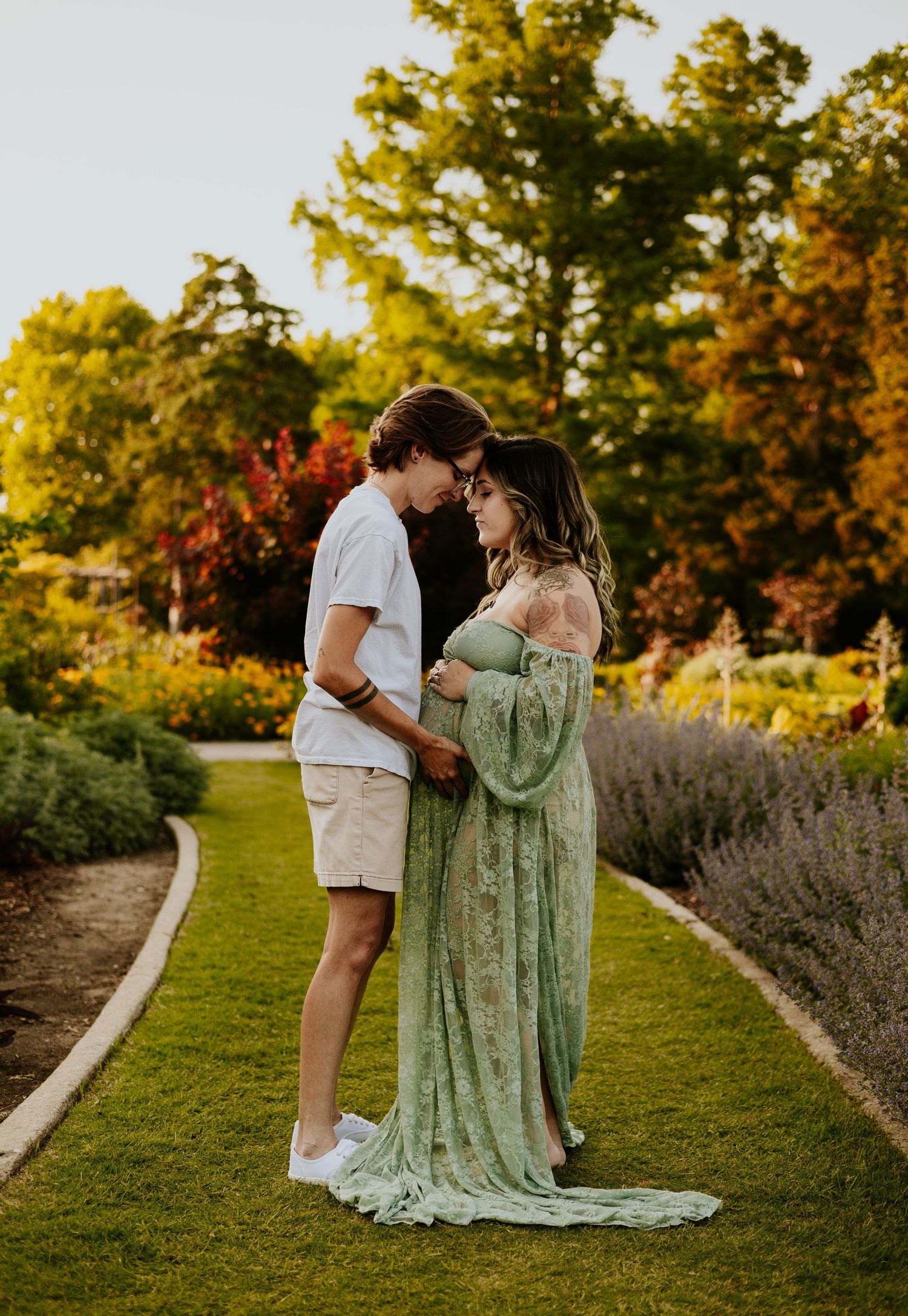 Couple sharing tender moment in garden at sunset, pregnant woman in green lace gown embraced by partner.