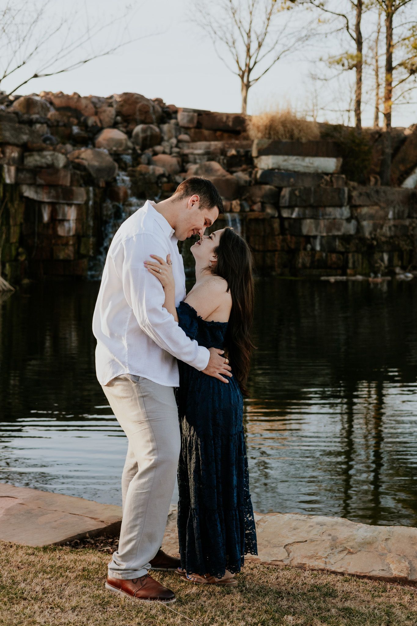 Couple embracing near rocky waterfall at sunset, woman in navy blue dress, man in white shirt.