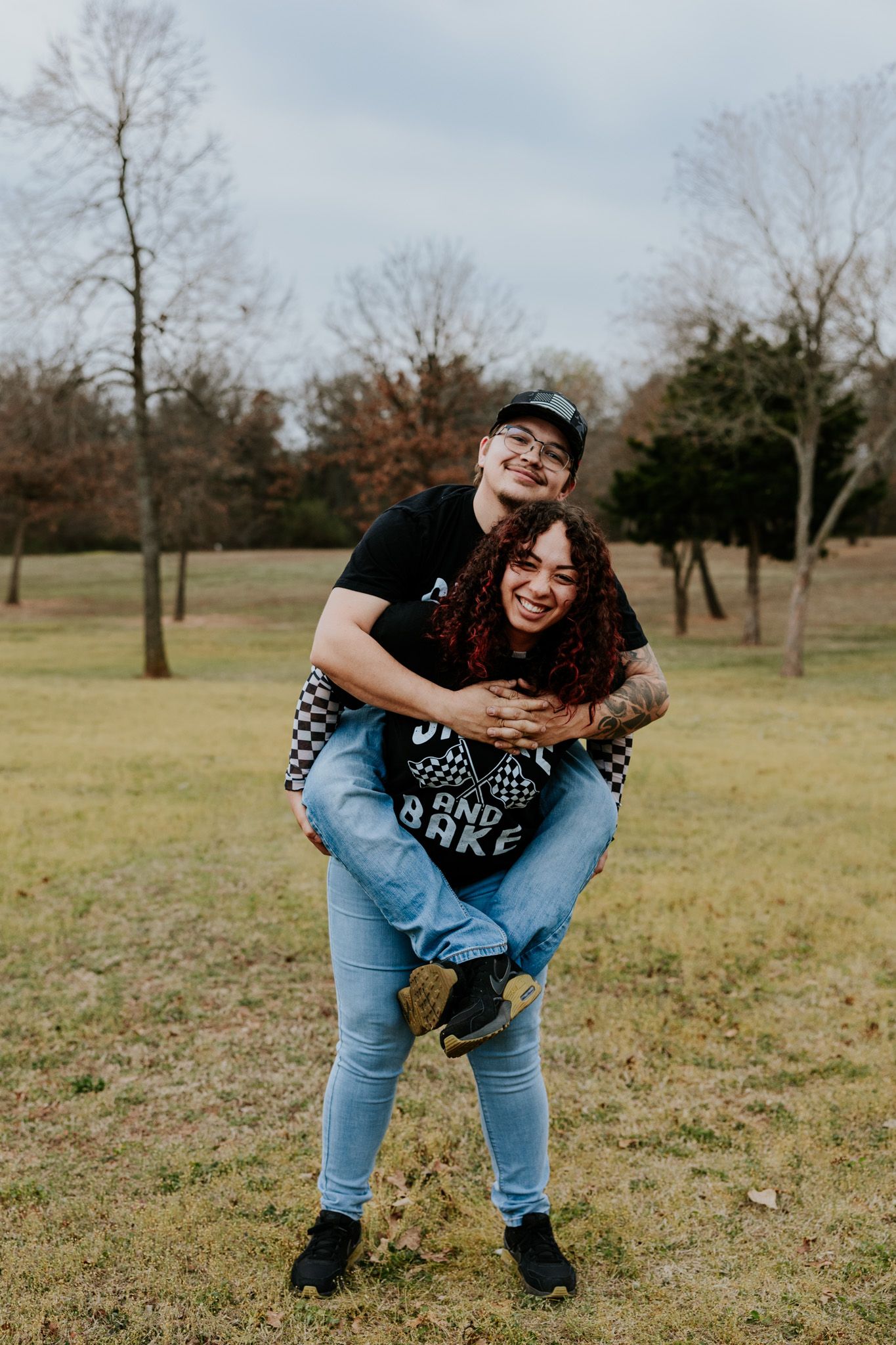 Man giving woman a piggyback ride while laughing in an open grassy park during couples photoshoot.