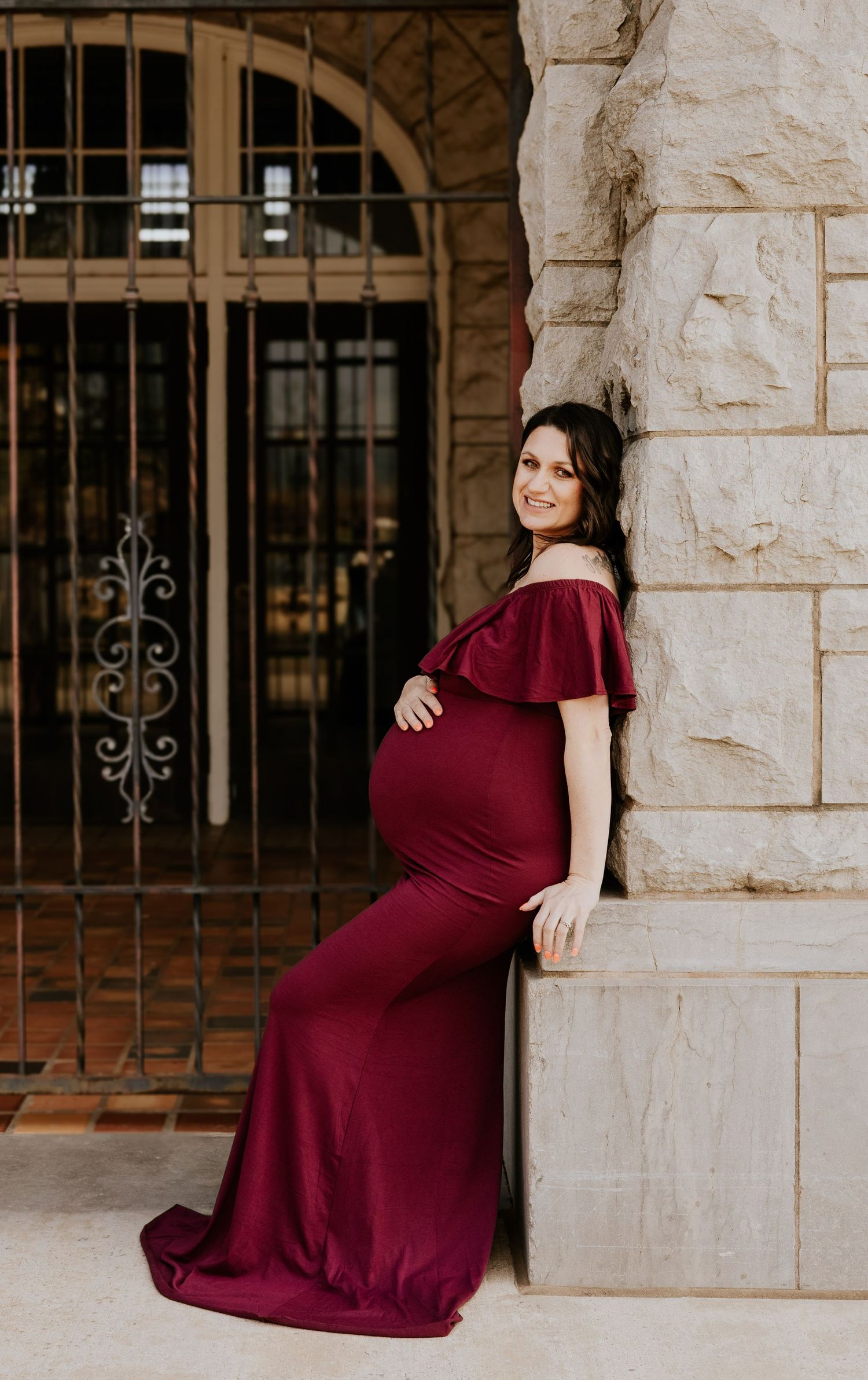 Smiling pregnant woman in dark red off-shoulder maternity dress leaning against stone pillar.