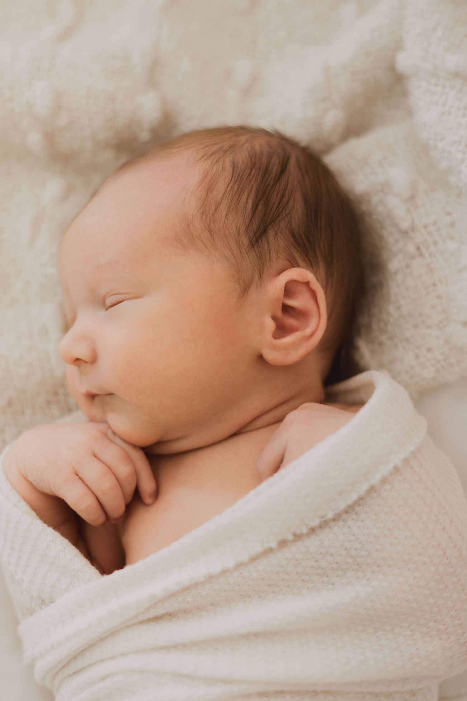 Sleeping newborn baby swaddled in a white knit wrap resting on a soft blanket.