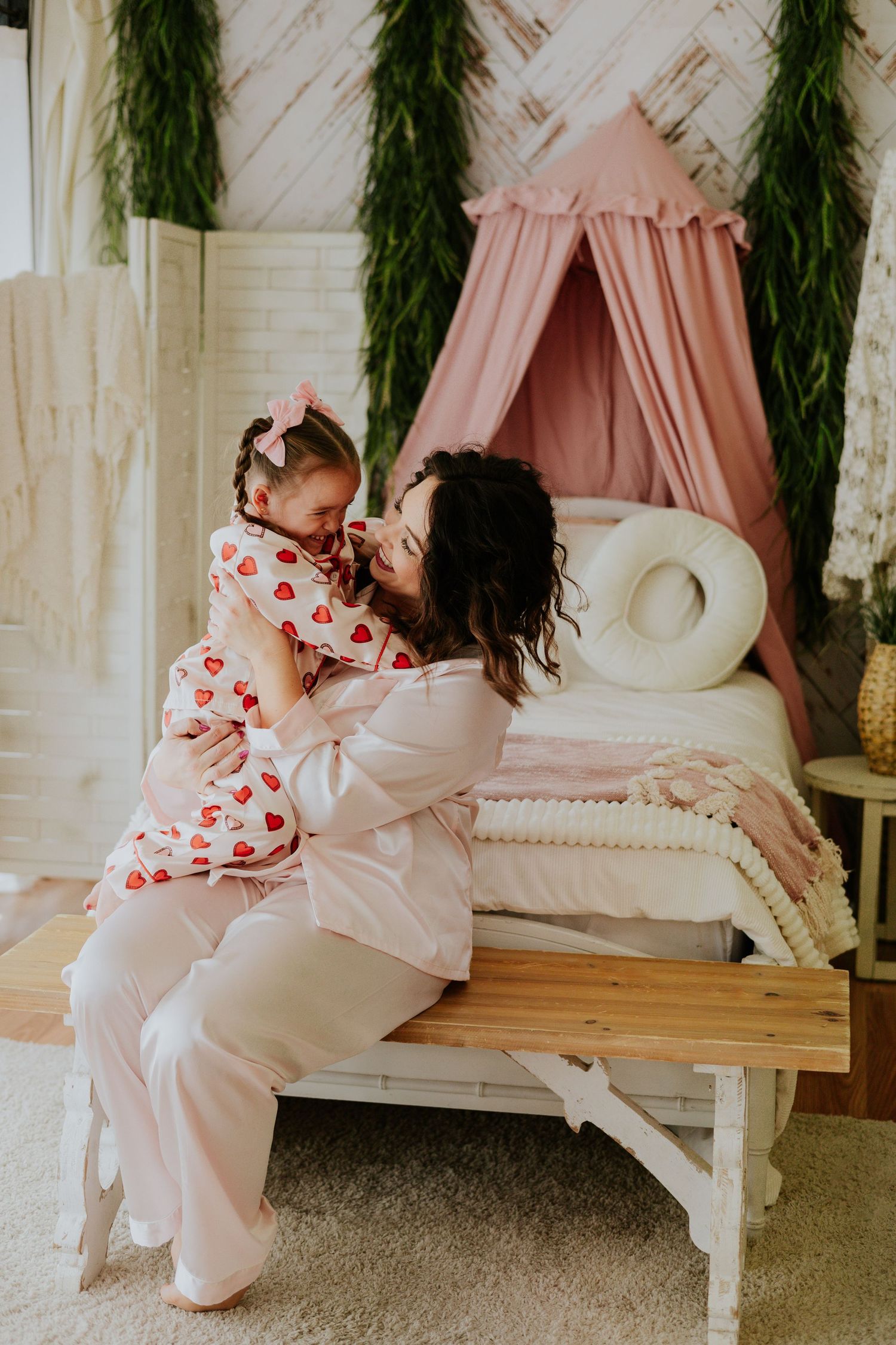 Pregnant mother in pink pajamas sits on bench hugging young daughter in heart-print pajamas indoors.