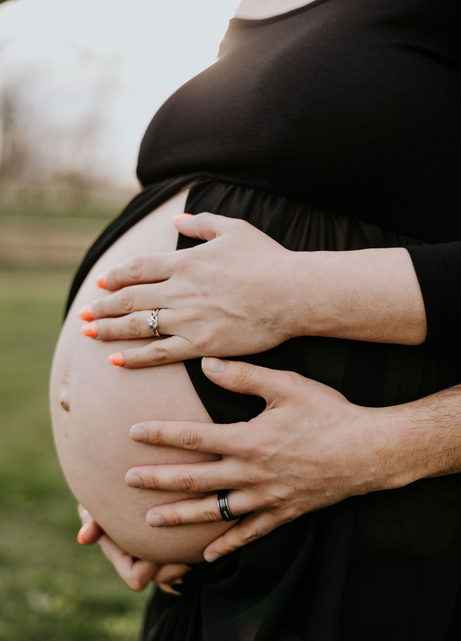 Close-up of pregnant belly with couple's hands, woman wearing orange nail polish and engagement ring.