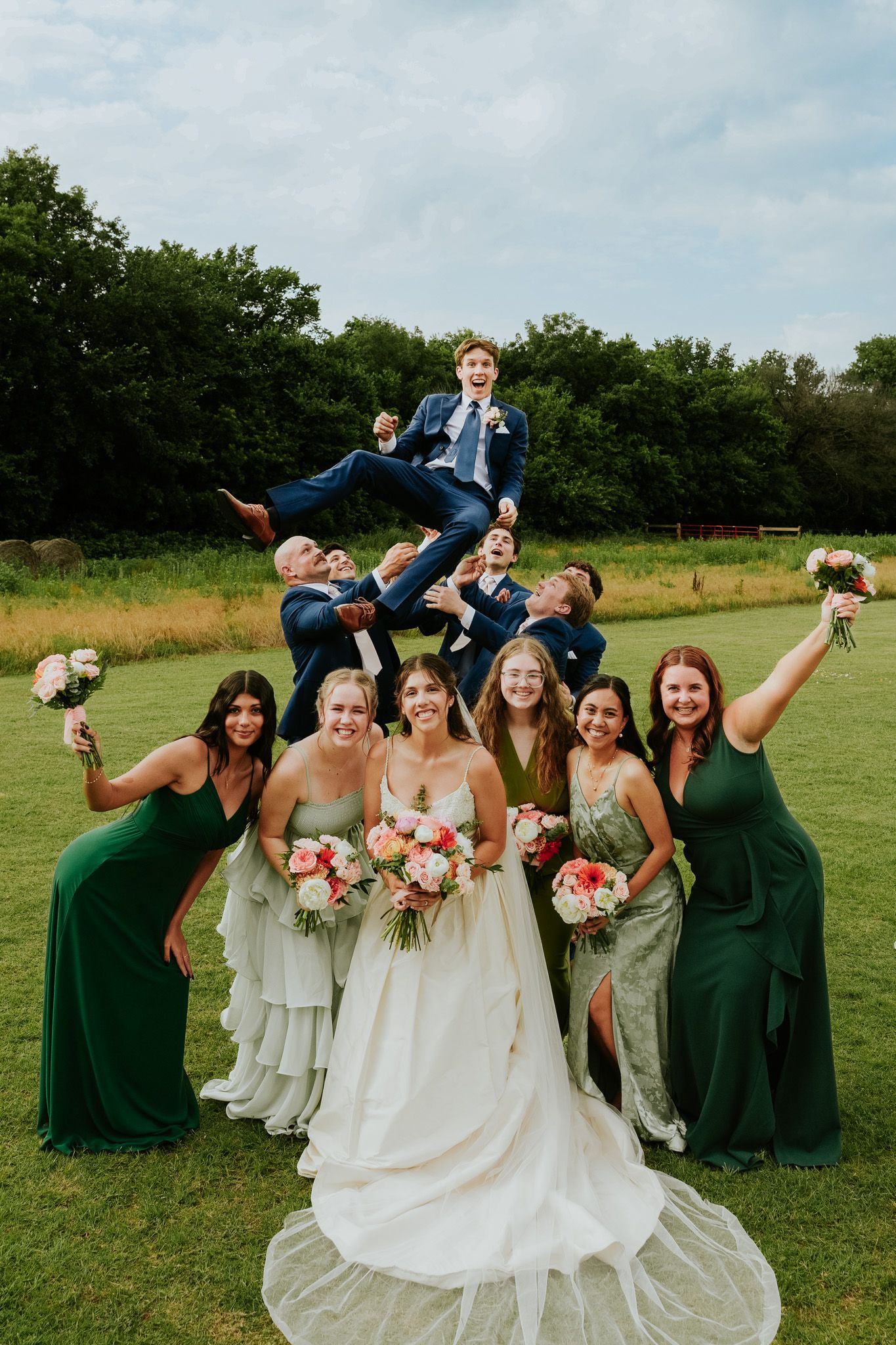 Wedding party celebrates outdoors, bridesmaids in green dresses hold bouquets as groomsmen toss groom in the air.