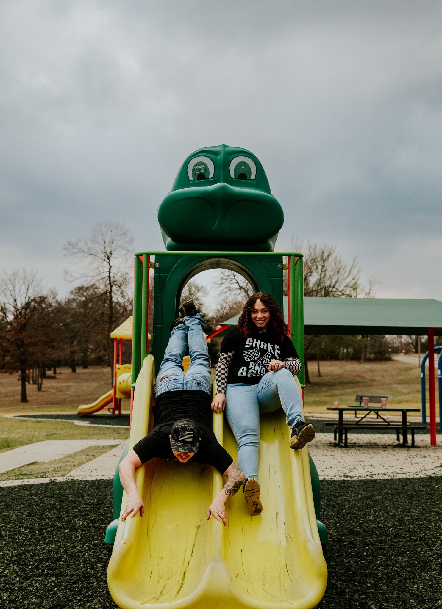 Fun couple sliding down frog-shaped playground slide together on cloudy day at outdoor park.