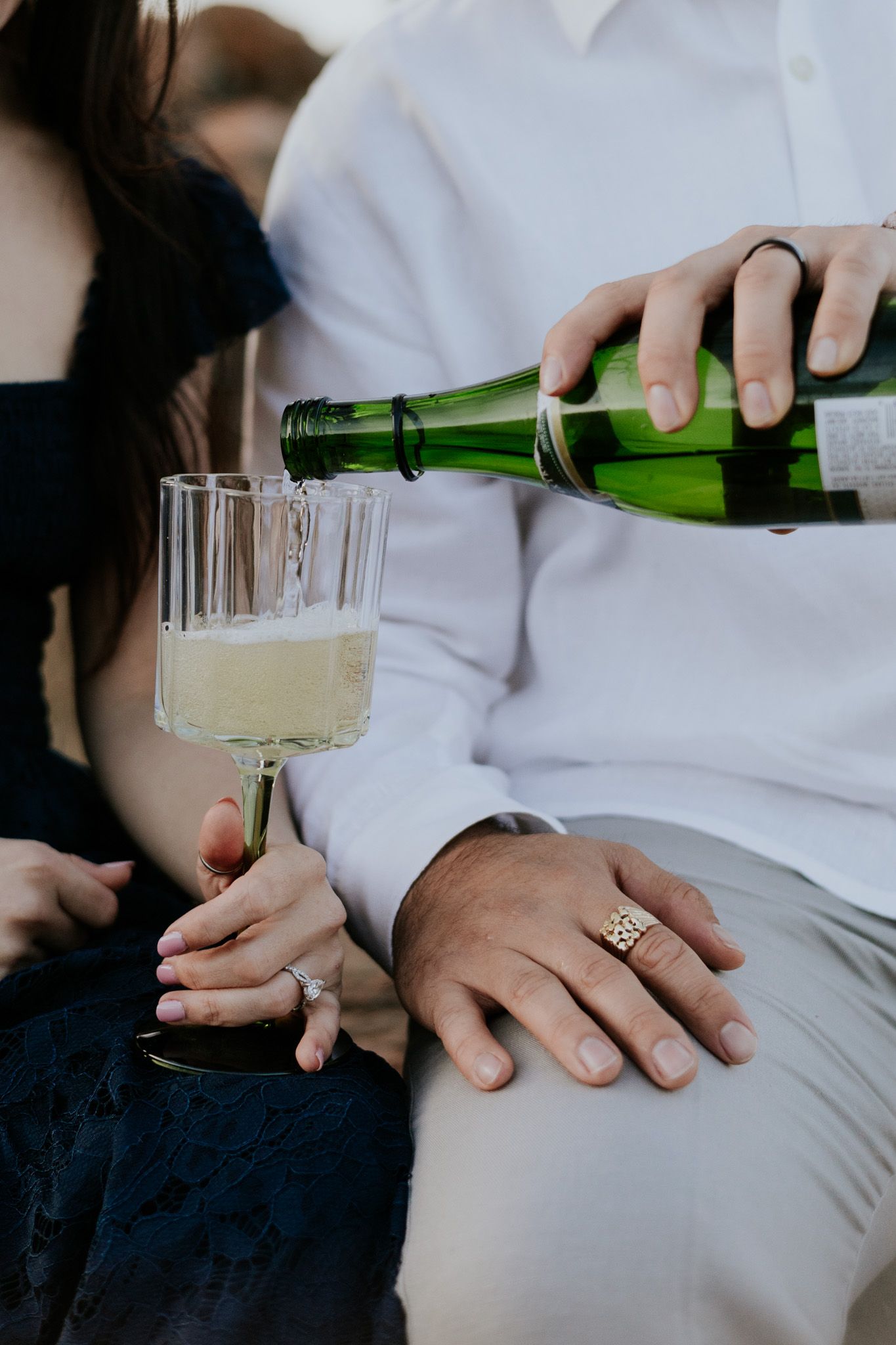 Close-up of man pouring champagne into glass held by woman wearing diamond engagement ring outdoors.