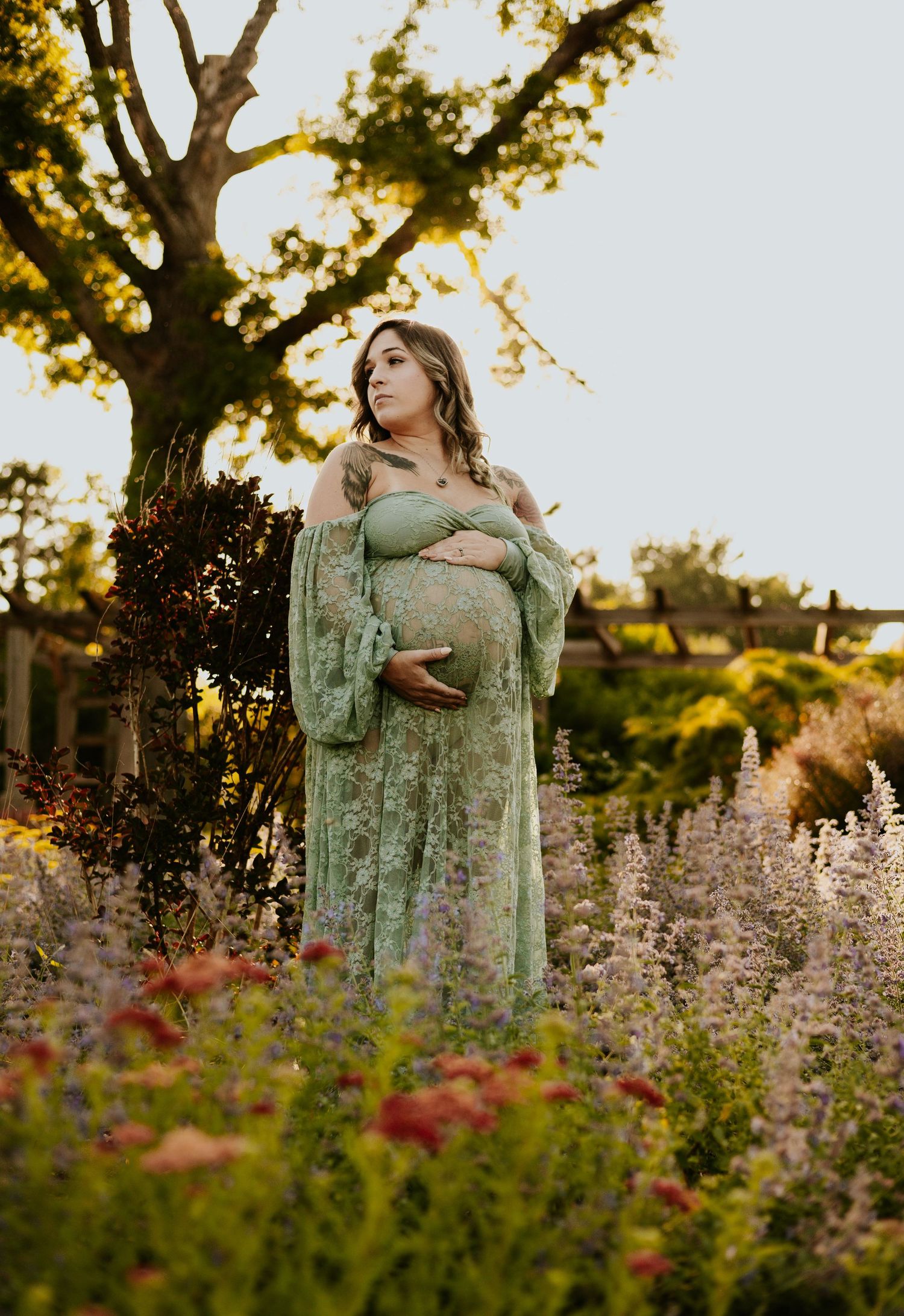 Pregnant woman in flowing green lace maternity gown standing among colorful wildflowers at golden hour sunset.