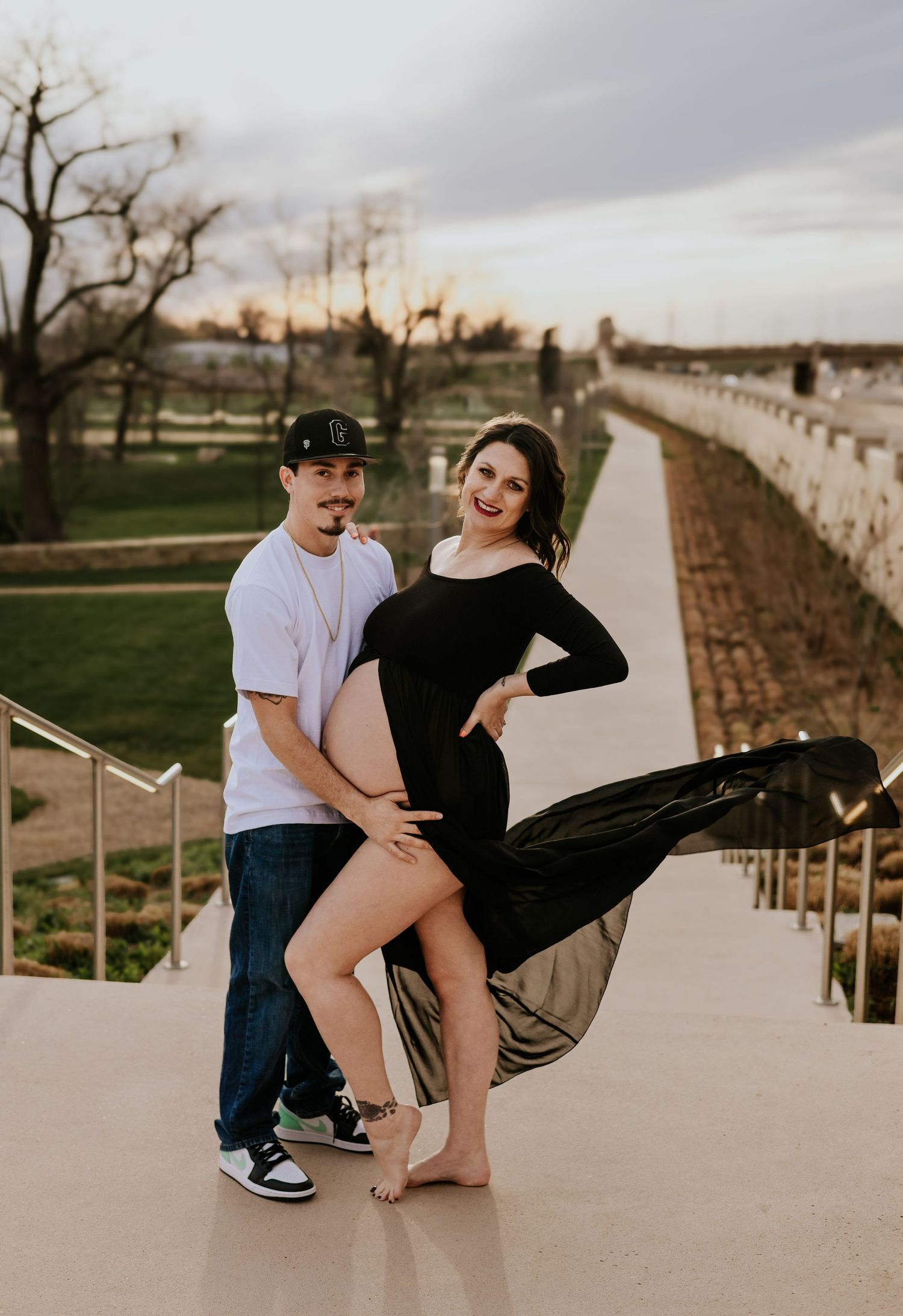 Couple poses together for maternity photo shoot outdoors at sunset, woman in black flowing gown.
