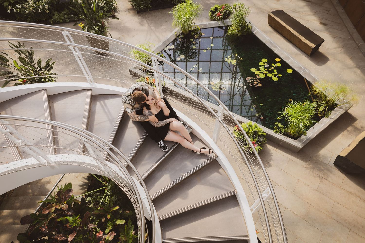 Aerial view of a couple embracing on a curved spiral staircase surrounded by lush greenery and a pond.