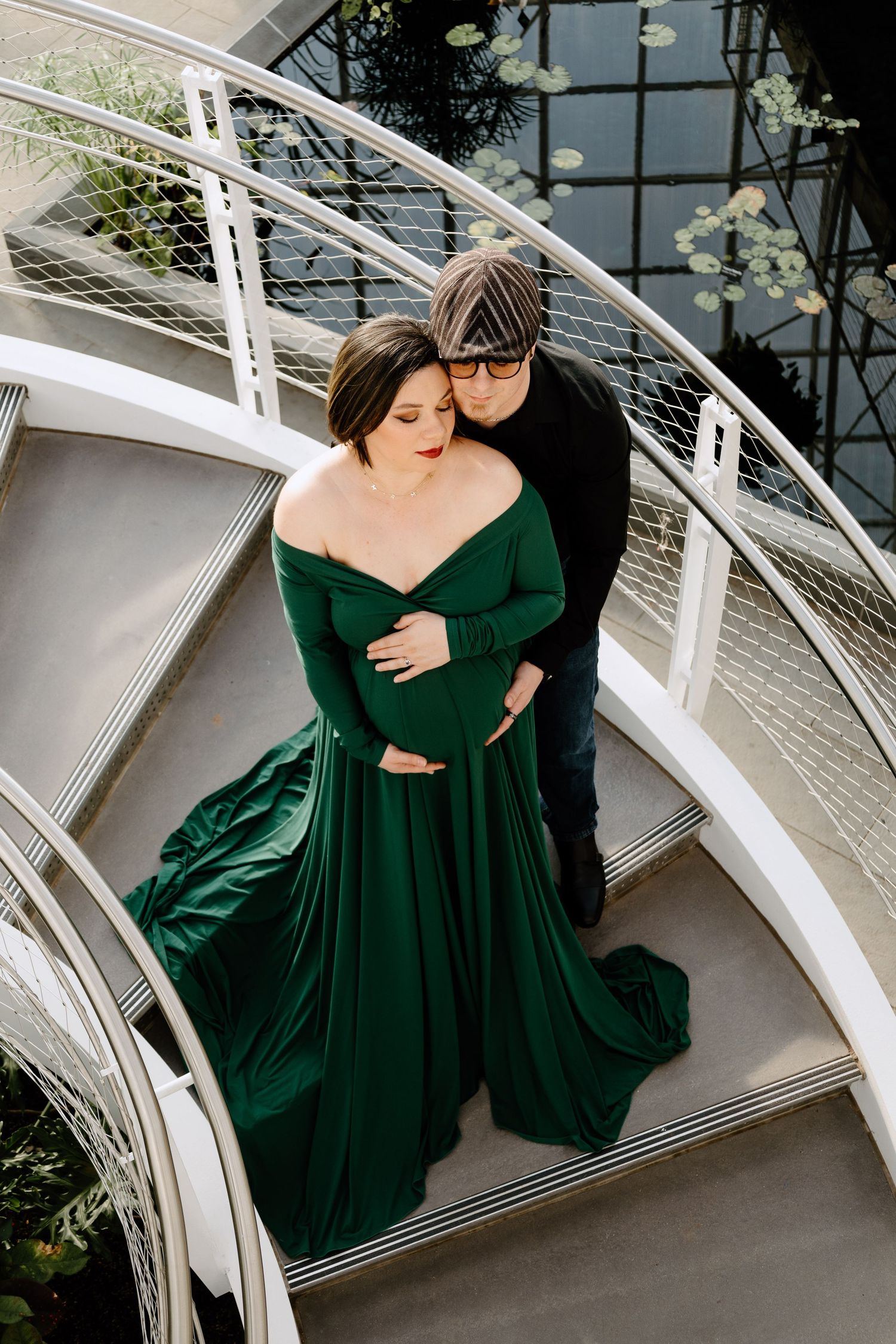 Couple embracing on spiral staircase, woman in stunning emerald maternity gown in a botanical garden.