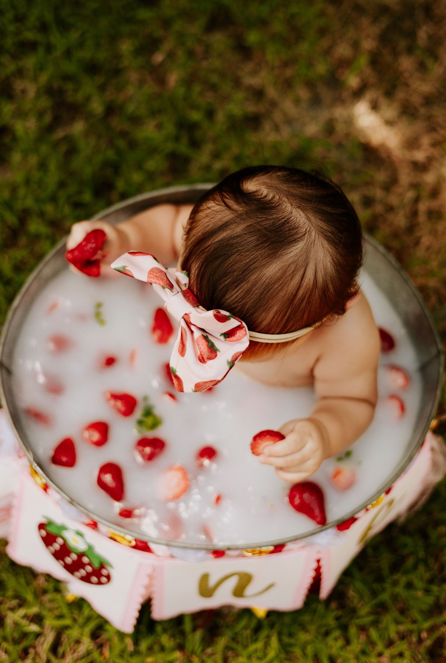 Overhead view of a baby girl with a strawberry bow headband sitting in a milk bath with fresh strawberries.