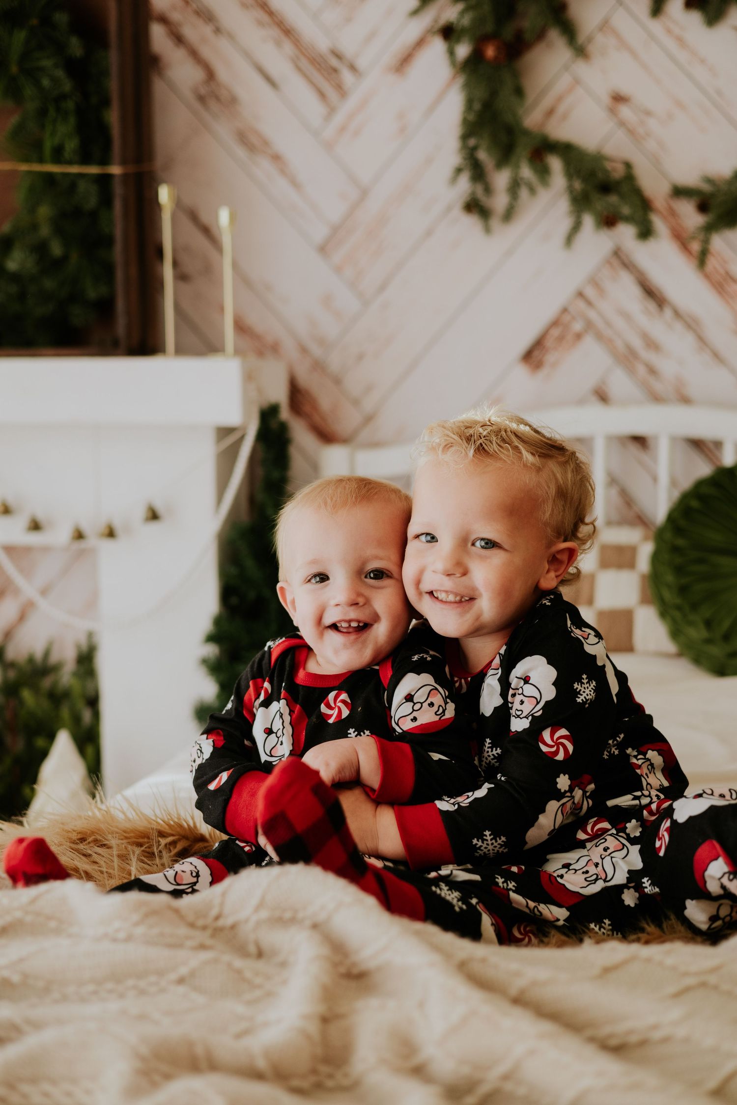 Two young brothers in matching Santa Claus Christmas pajamas hugging and smiling on a cozy bed.