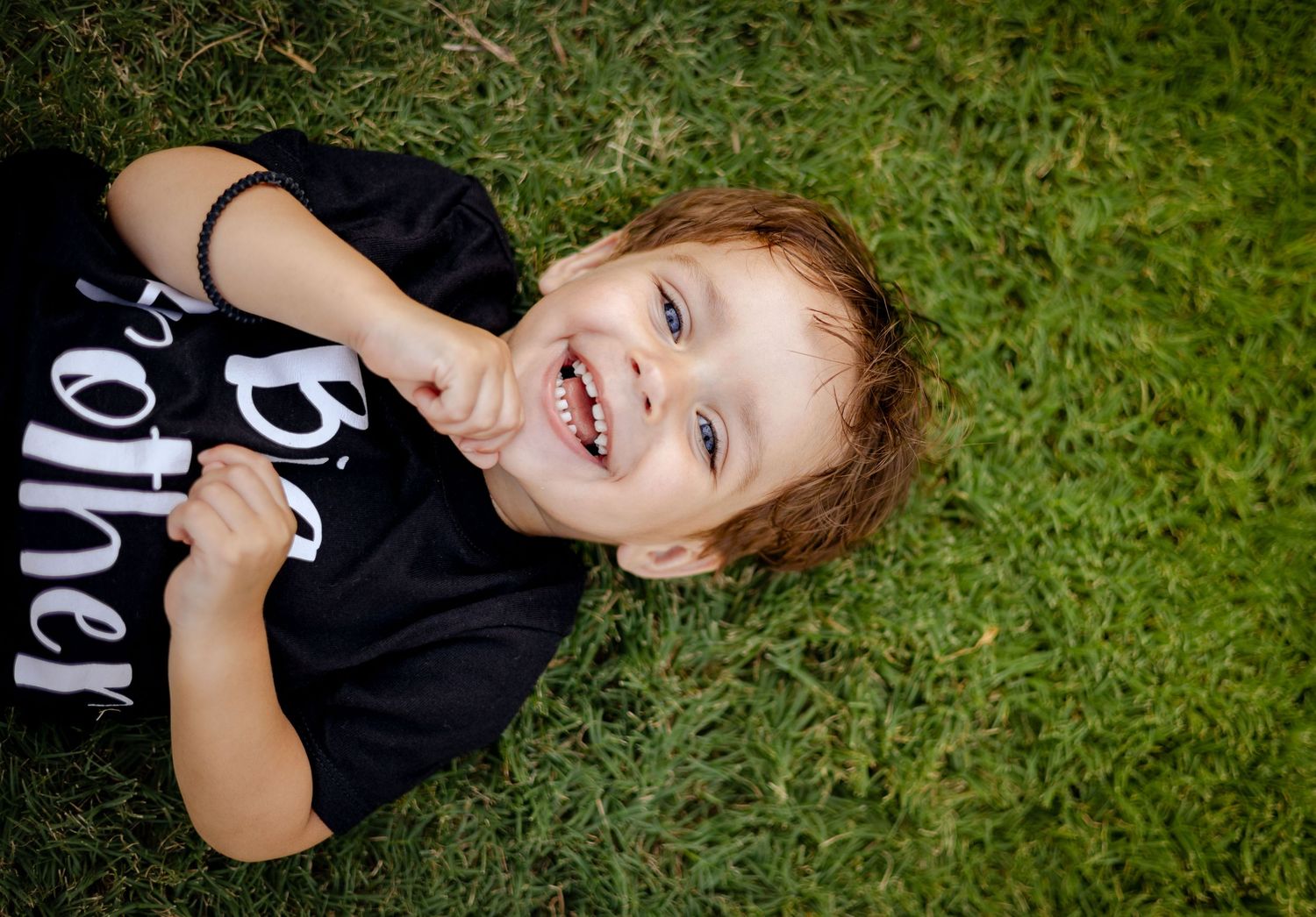 Happy toddler in Big Brother shirt laughing while lying on green grass.