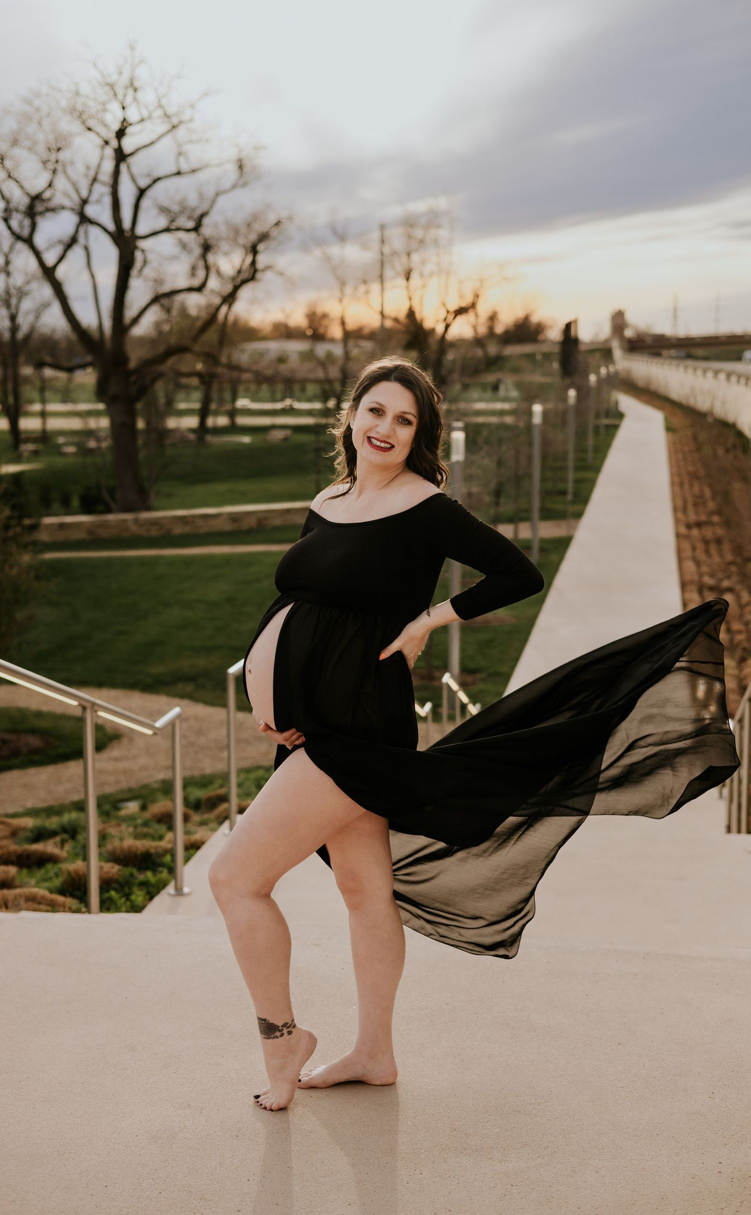 Pregnant woman in flowing black maternity gown poses barefoot on outdoor walkway at sunset.