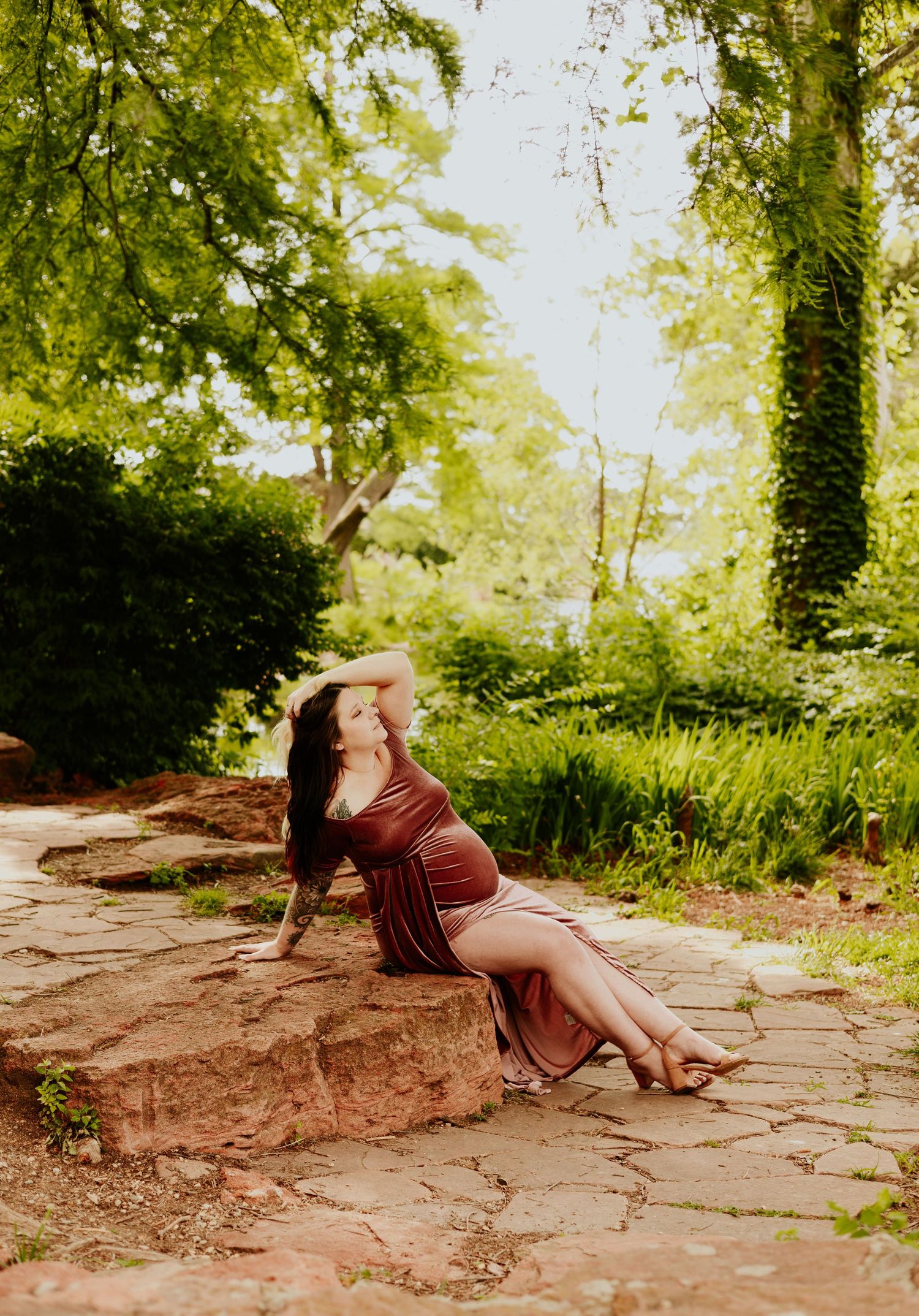 Pregnant woman in a rust velvet gown posing gracefully on stone steps in a lush garden.