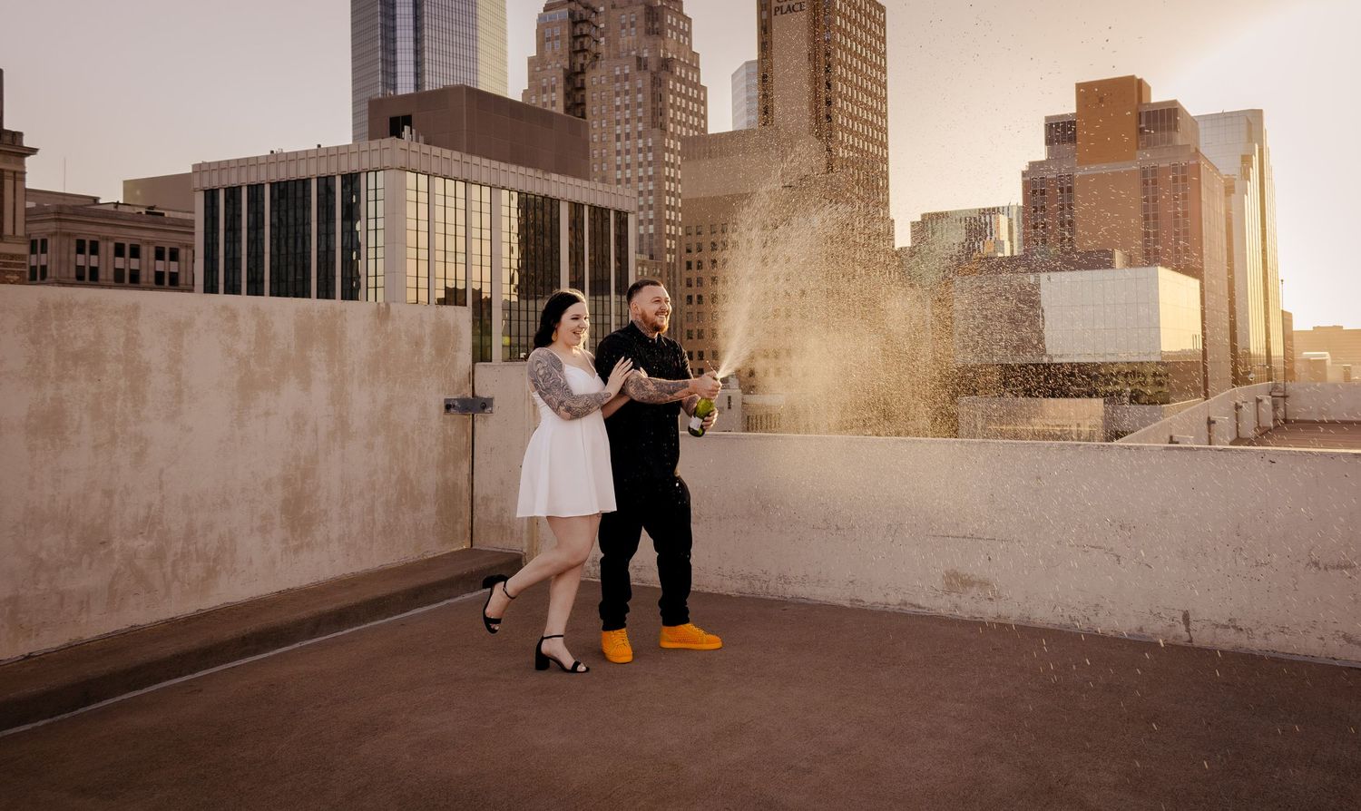Couple popping champagne on rooftop with city skyline at sunset.