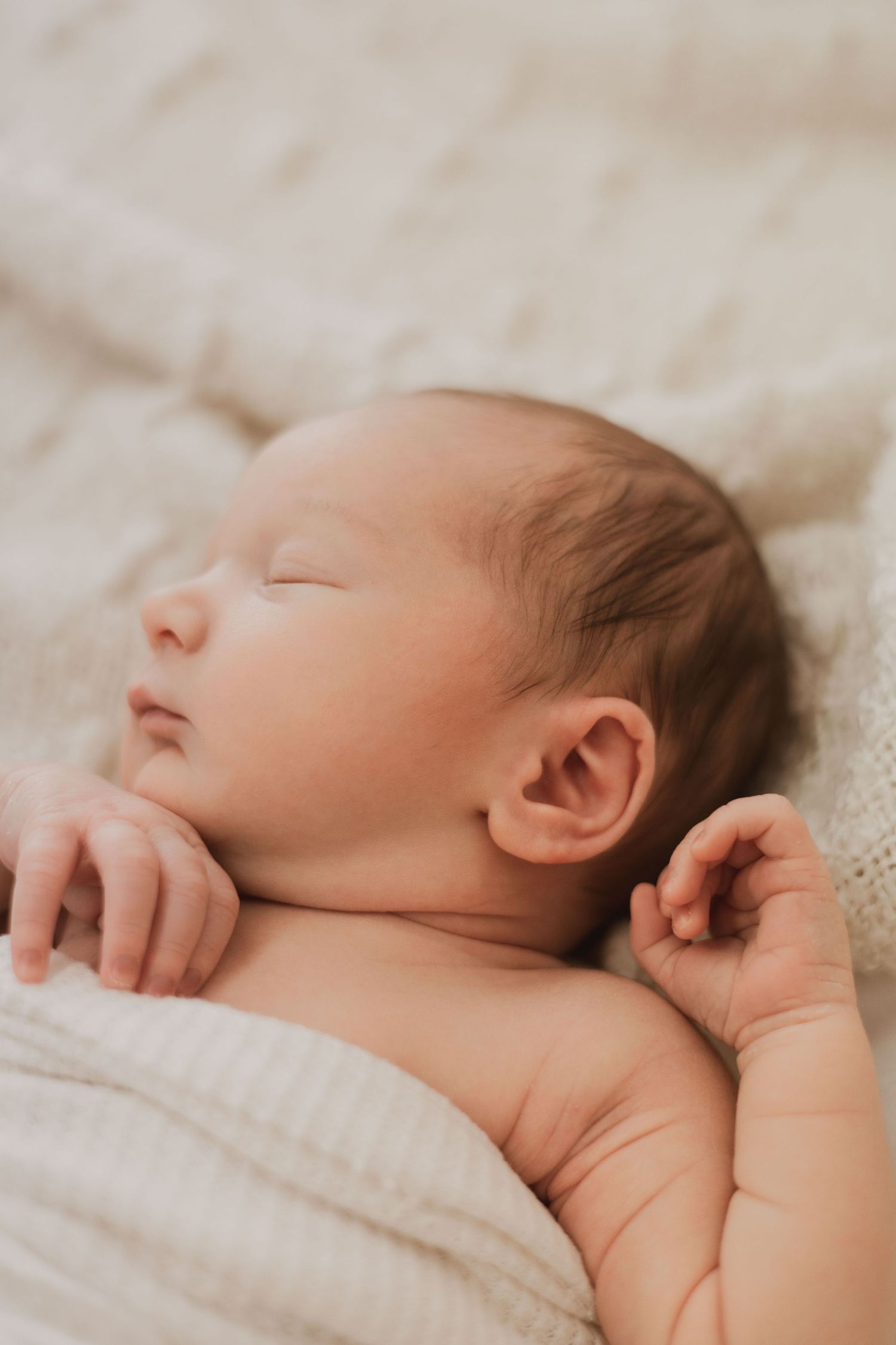 Peaceful newborn baby sleeping on a white knit blanket with tiny hands raised.