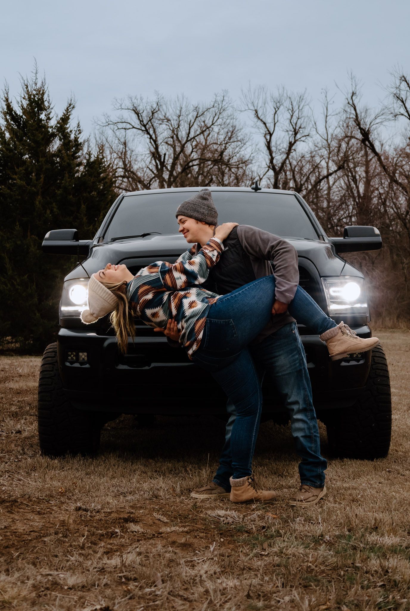 Couple laughing and dancing in front of a lifted RAM truck with headlights on at dusk.