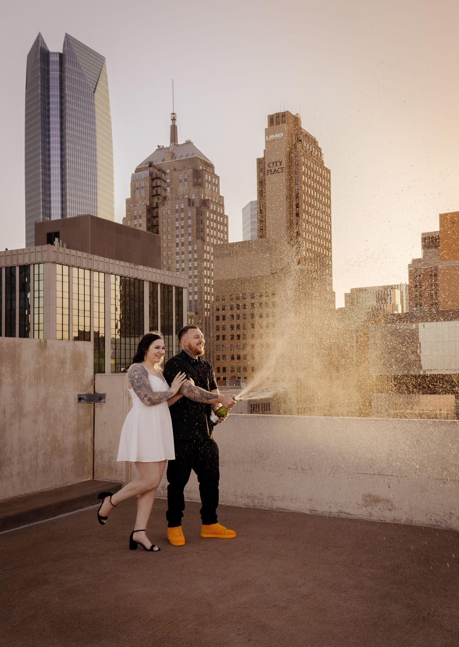 Couple celebrating with champagne spray on urban rooftop at sunset.