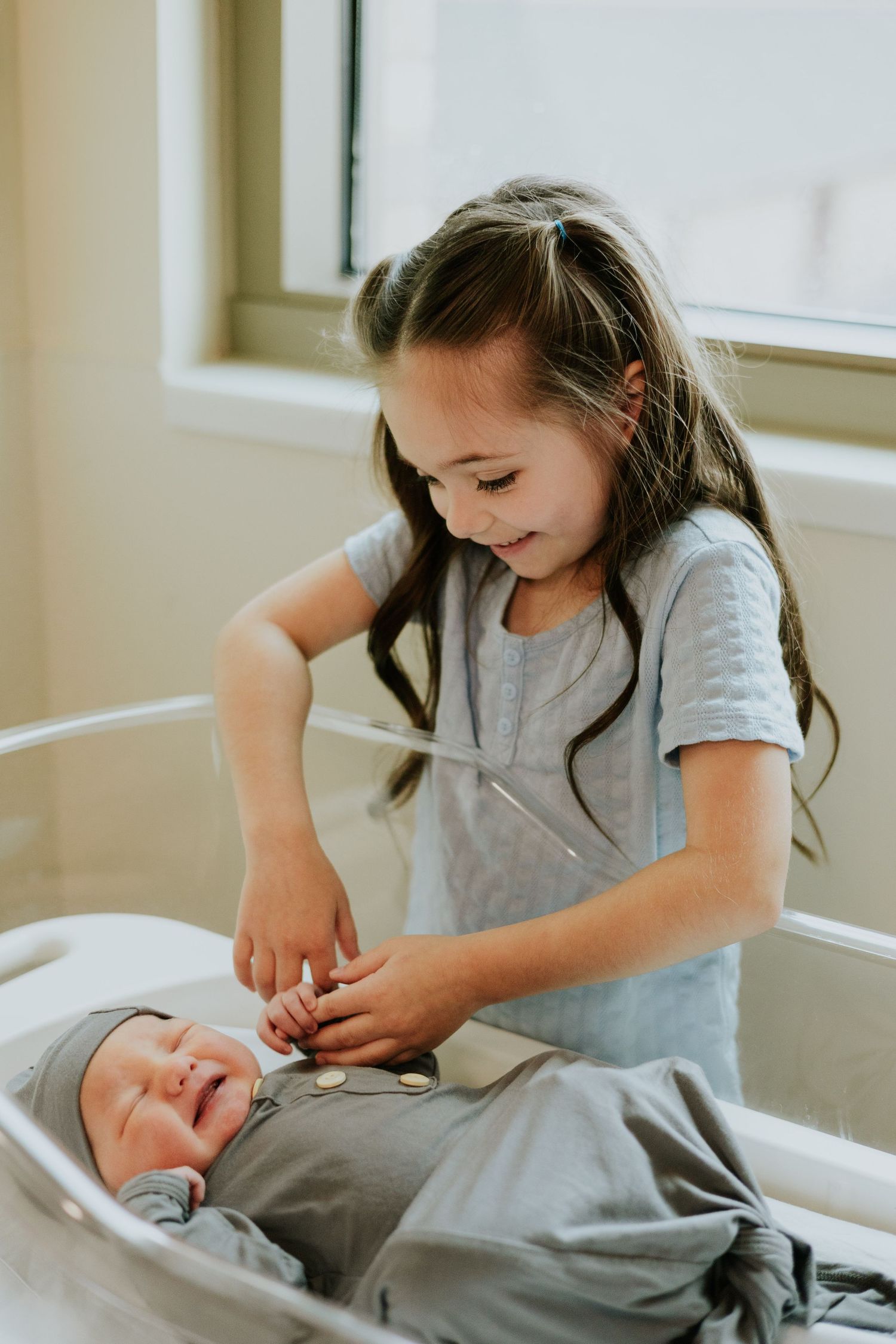 Young girl with long hair smiles at newborn baby in hospital bassinet, touching tiny hands gently.