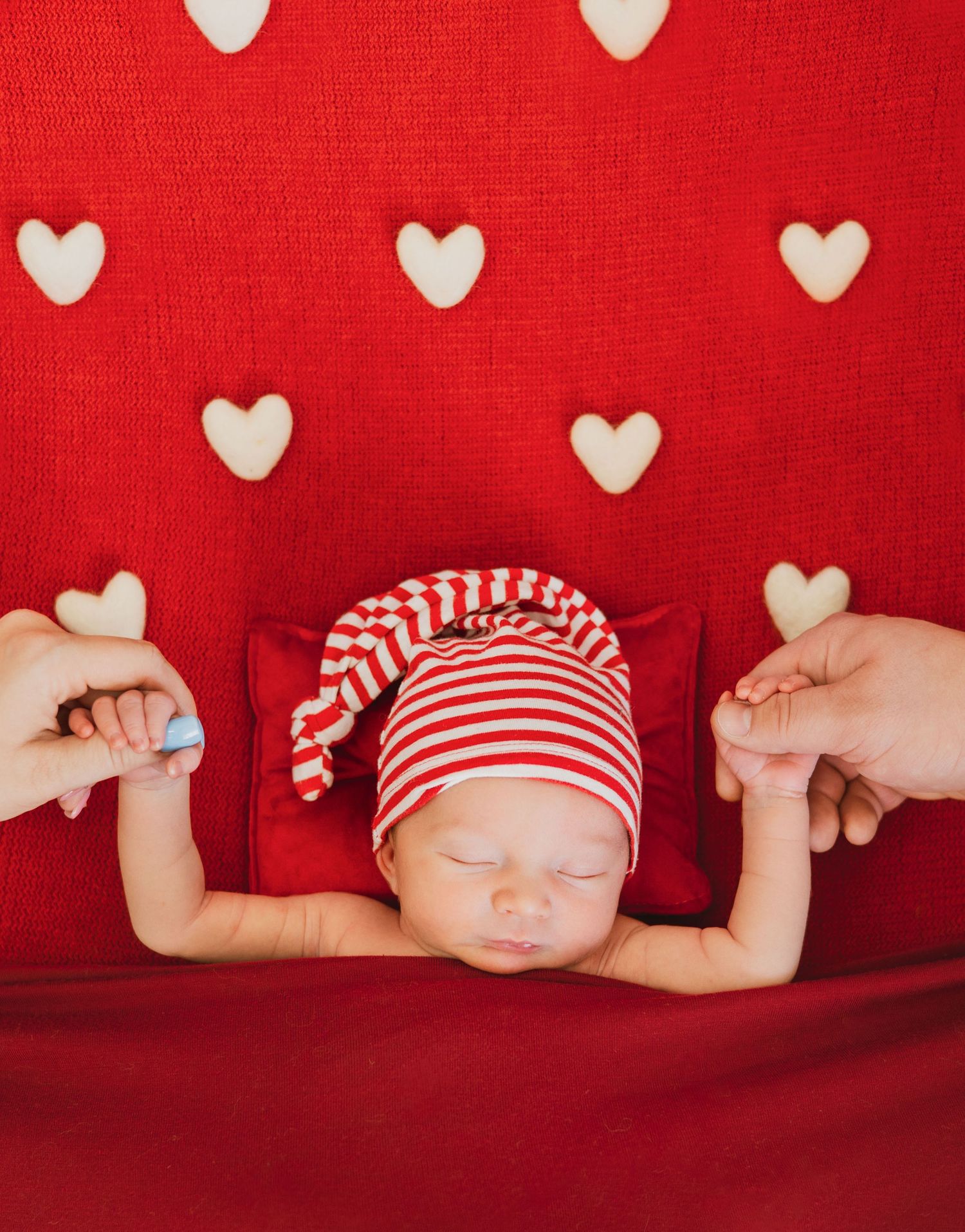Sleeping newborn in striped hat holding parents fingers on festive red heart-patterned Valentine background.