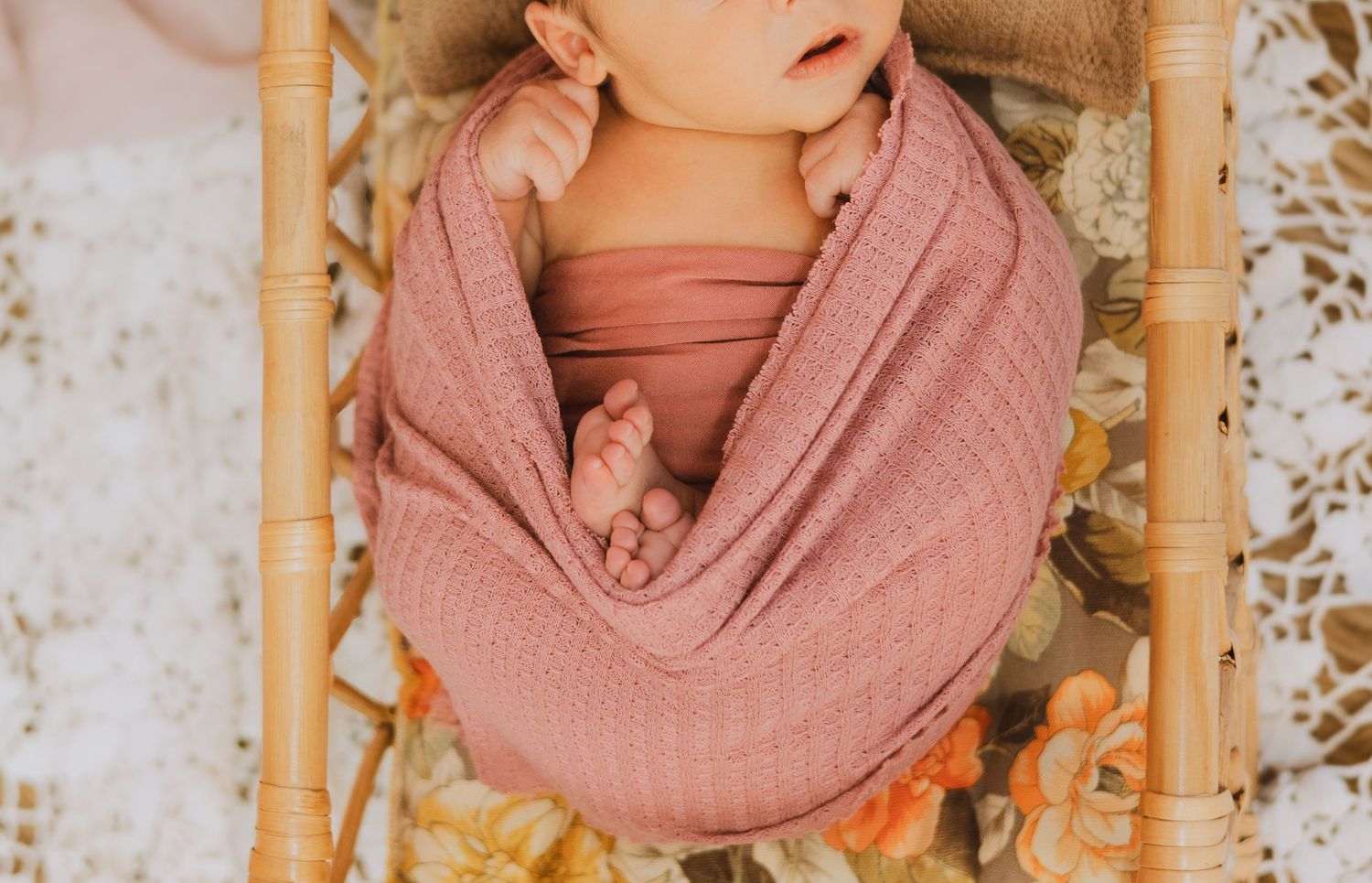 Newborn baby swaddled in pink wrap curled up in small wooden rattan bassinet.