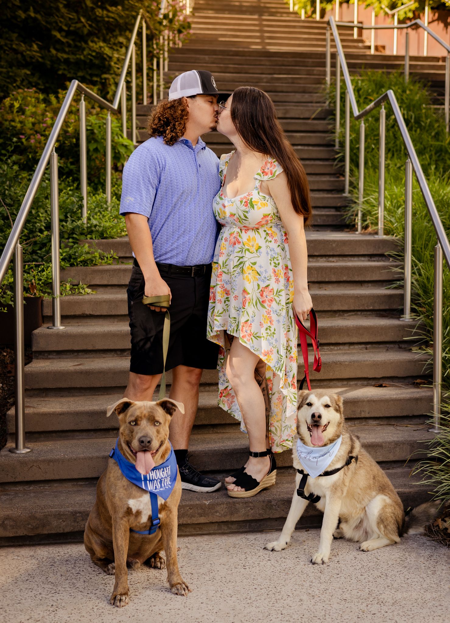 Couple kissing on steps with two dogs wearing bandanas outdoors.