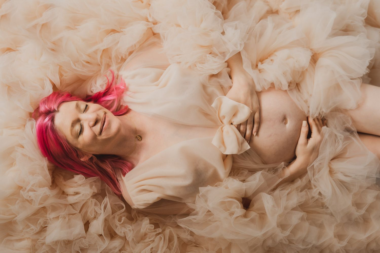 Pregnant woman with pink hair lying serenely in champagne tulle gown gently holding her baby bump.