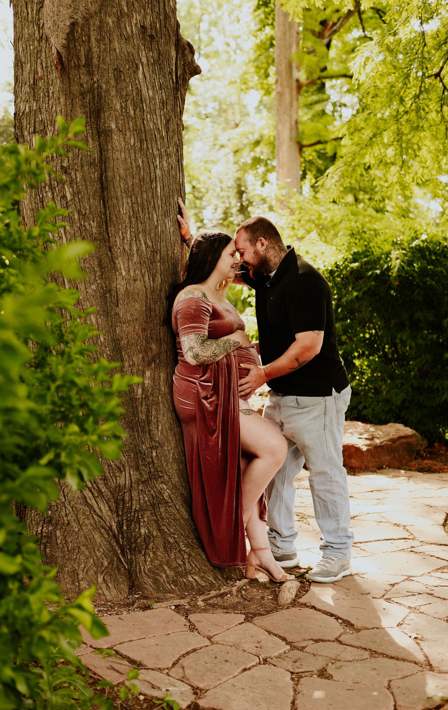 Pregnant couple sharing a tender moment leaning against a large tree in a lush garden, woman in elegant rust velvet gown.