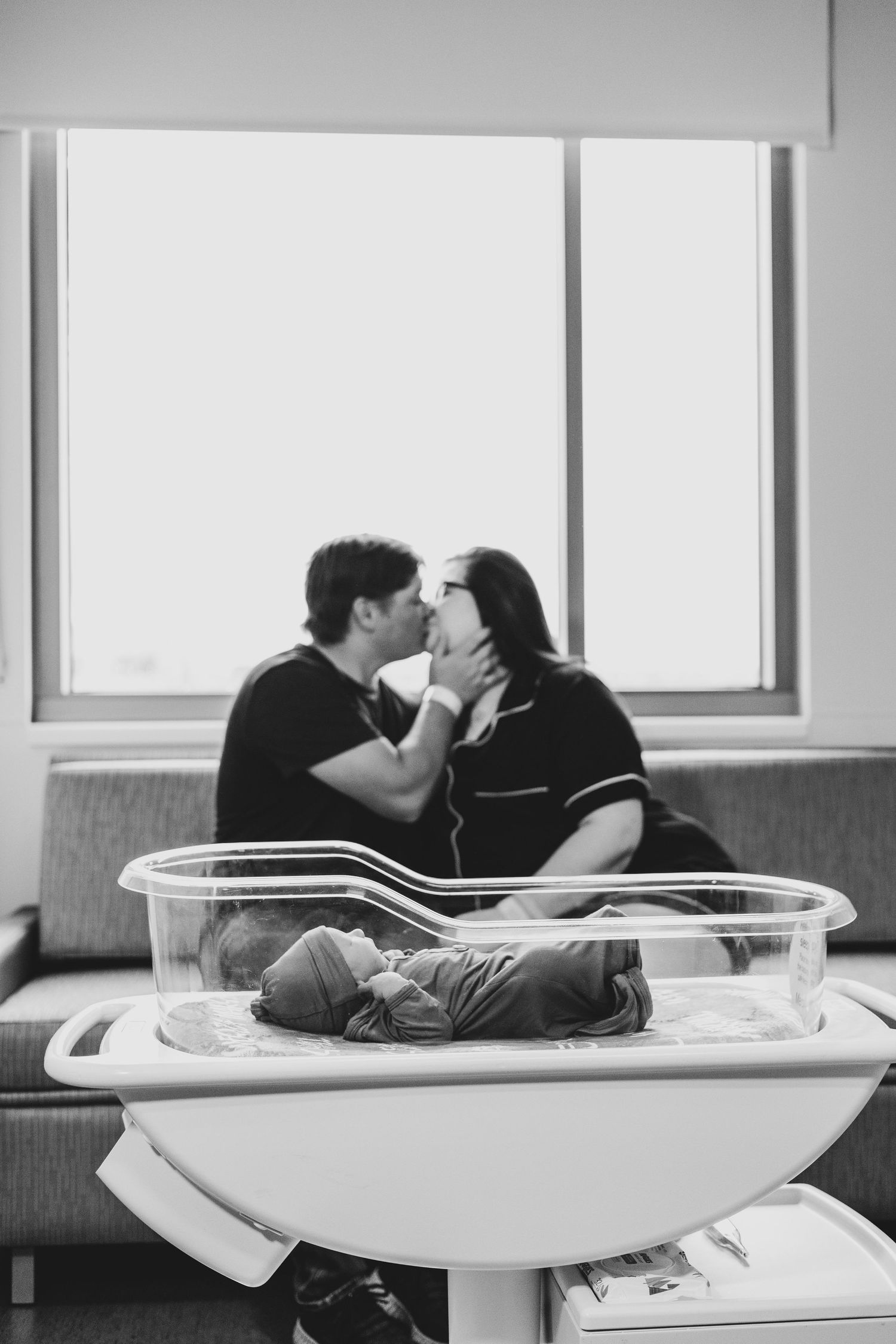 New parents kissing behind hospital bassinet with sleeping newborn in black and white photo.