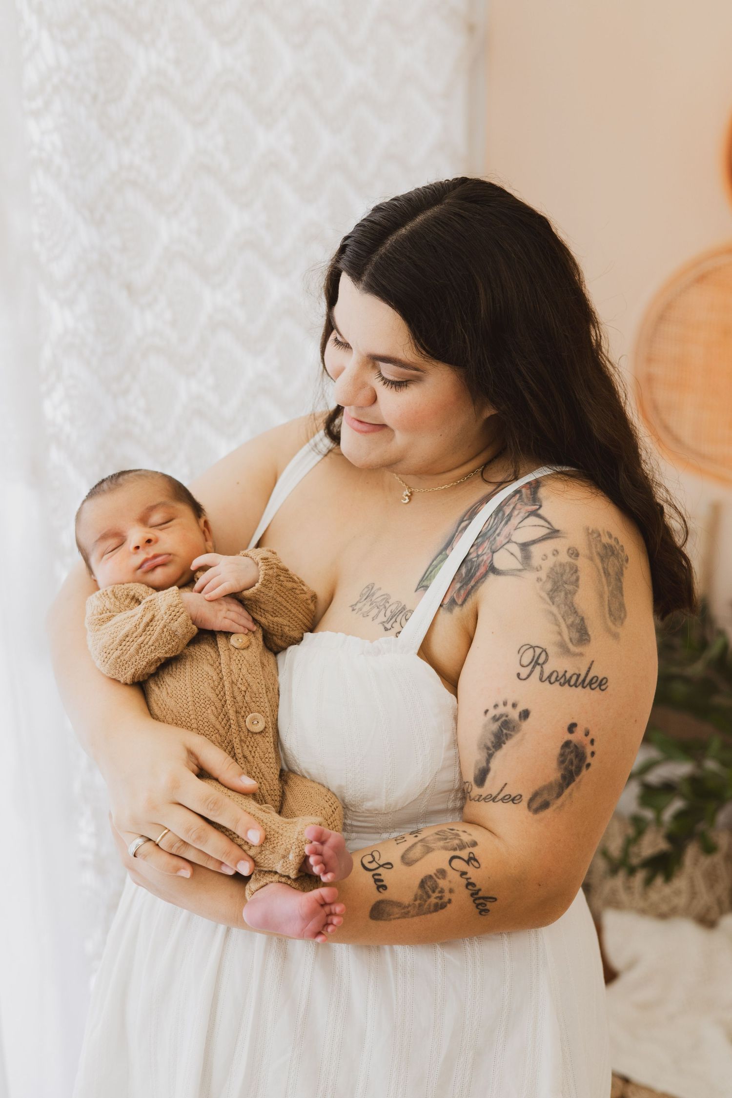 Tattooed mother in white dress tenderly holding sleeping newborn in brown knit outfit.