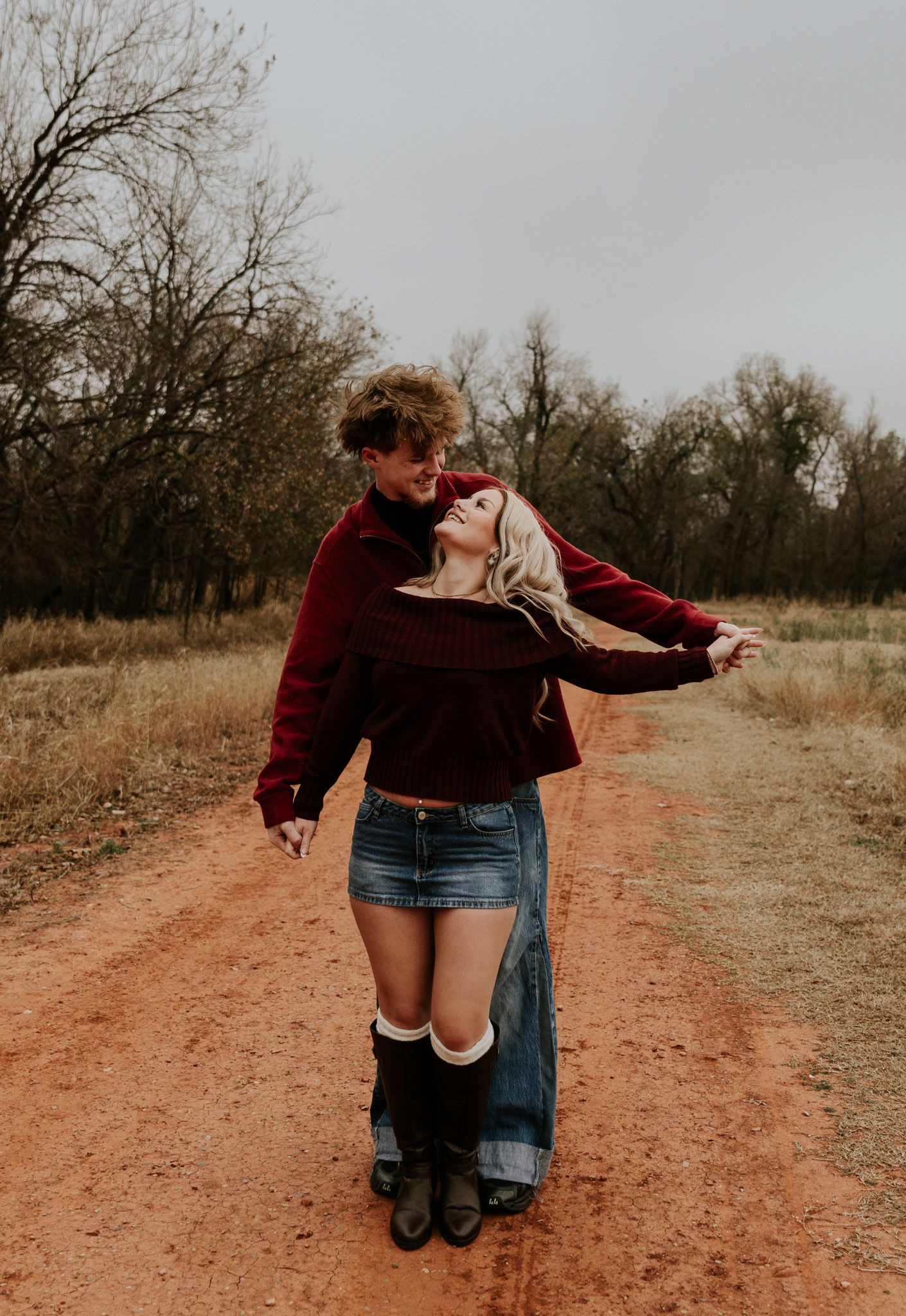 Couple embracing on a red dirt path surrounded by autumn trees, woman in denim skirt and knee-high boots.