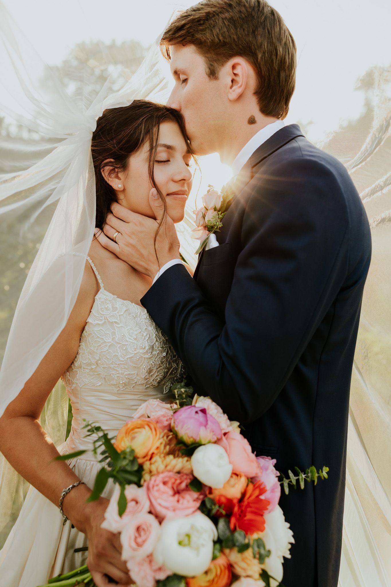 Groom in navy suit tenderly kisses bride in white lace gown under veil, holding colorful bouquet in golden sunlight.