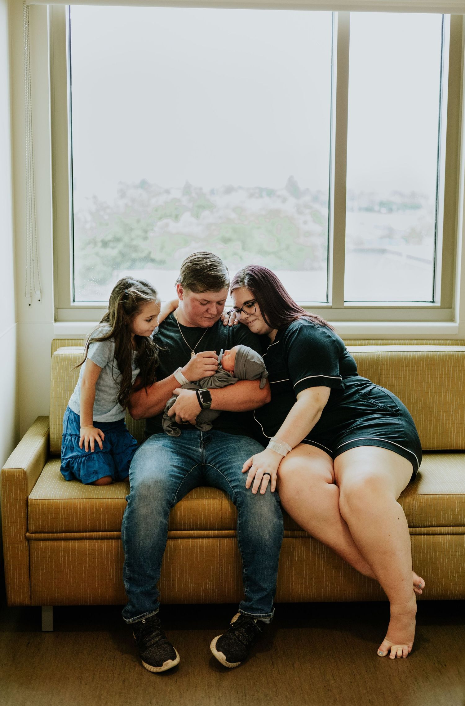 New parents and young daughter sitting on couch lovingly gazing at newborn baby in hospital.