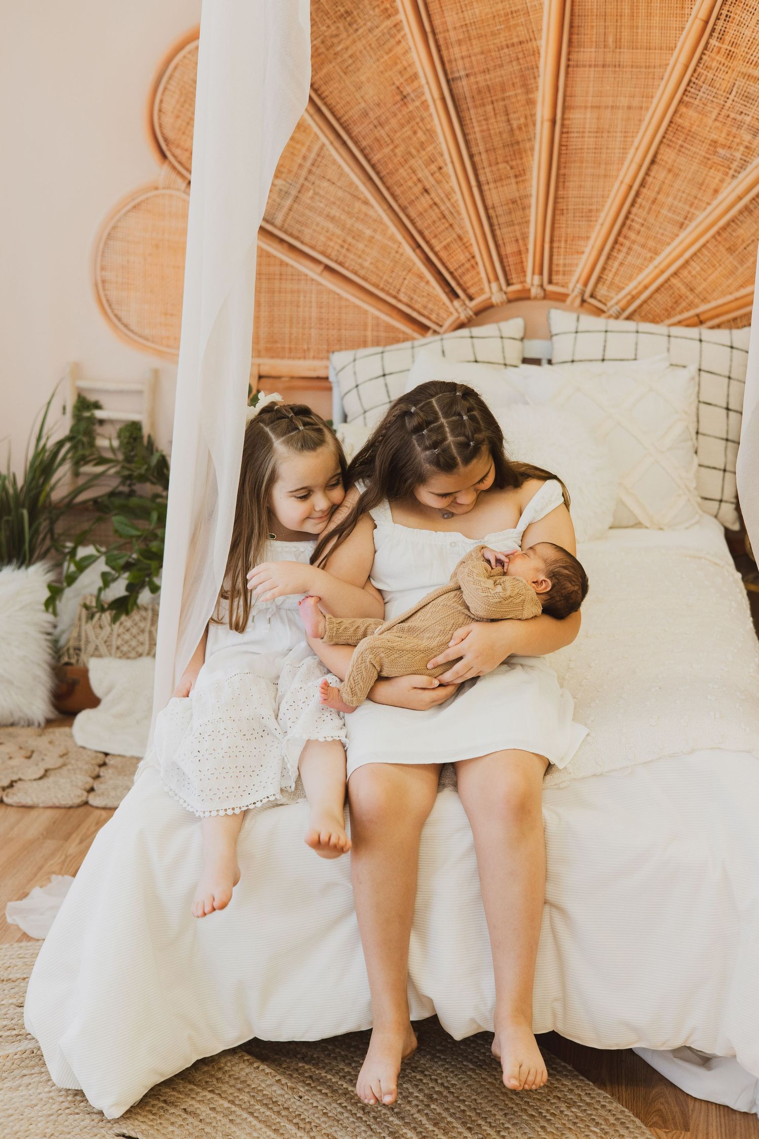 Two young girls in white dresses sitting on boho bed, gently holding sleeping newborn baby.
