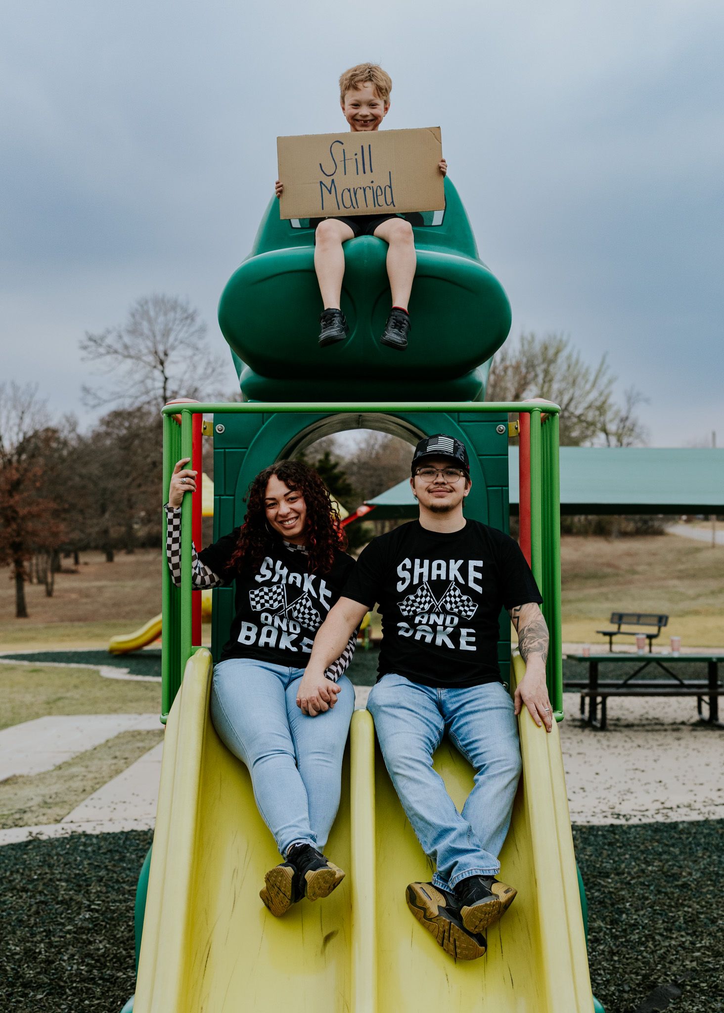 Couple in matching Shake and Bake shirts sitting on playground slide with child holding Still Married sign.