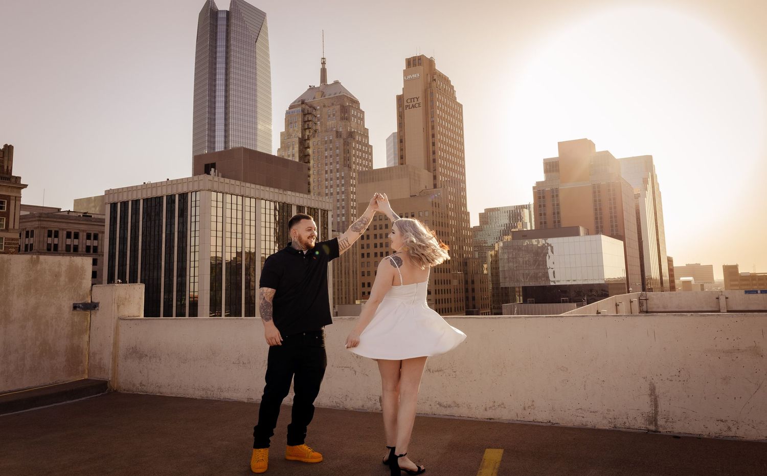 Man spinning woman in white dress on rooftop at sunset.