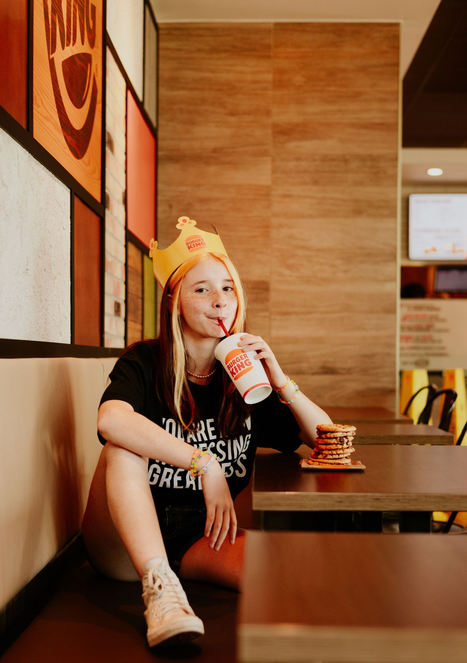 Teen girl in Burger King crown hat sipping drink while sitting in booth.