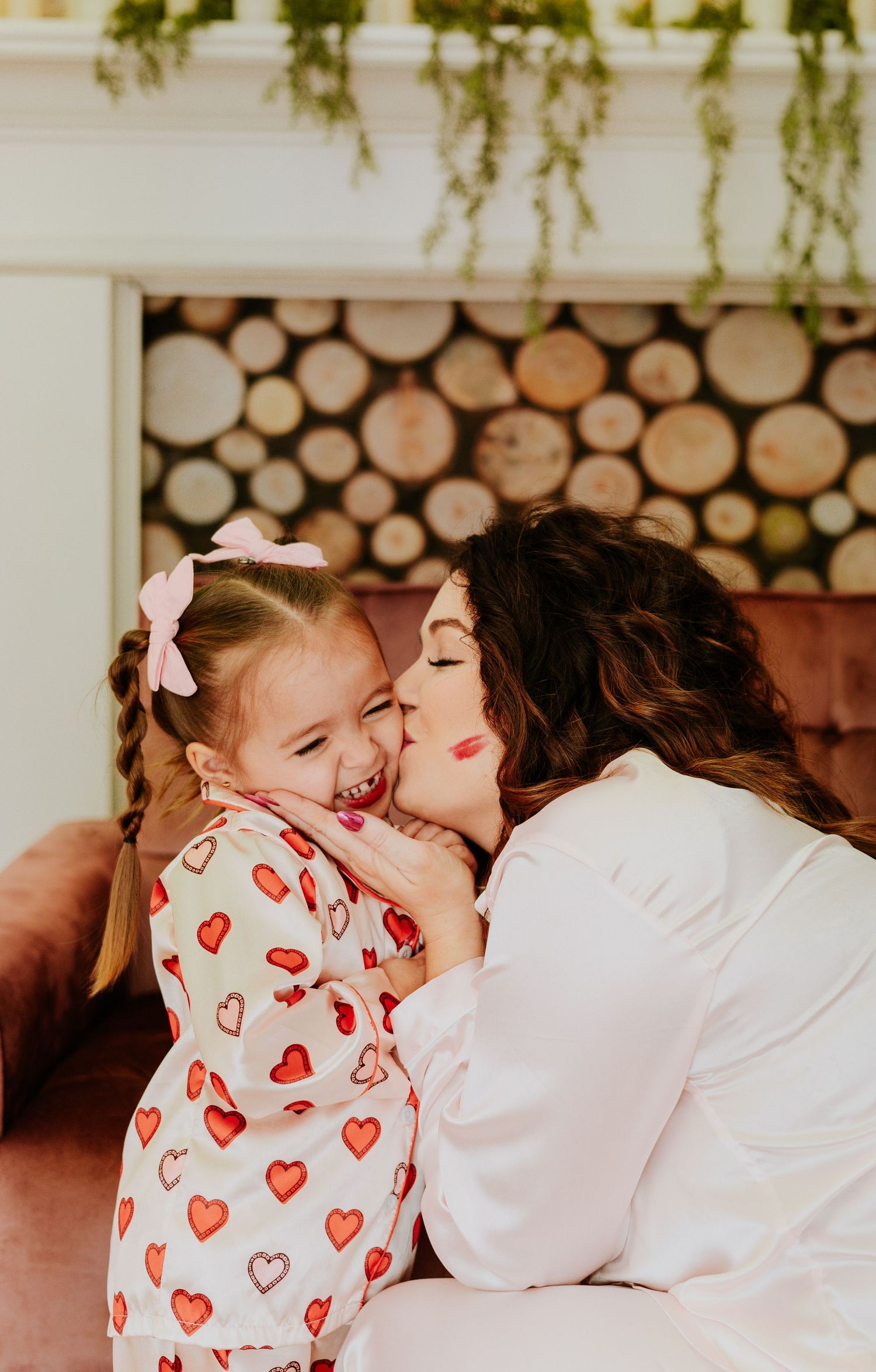 Mother lovingly kisses smiling young daughter wearing heart-print pajamas and pink bow in her hair.
