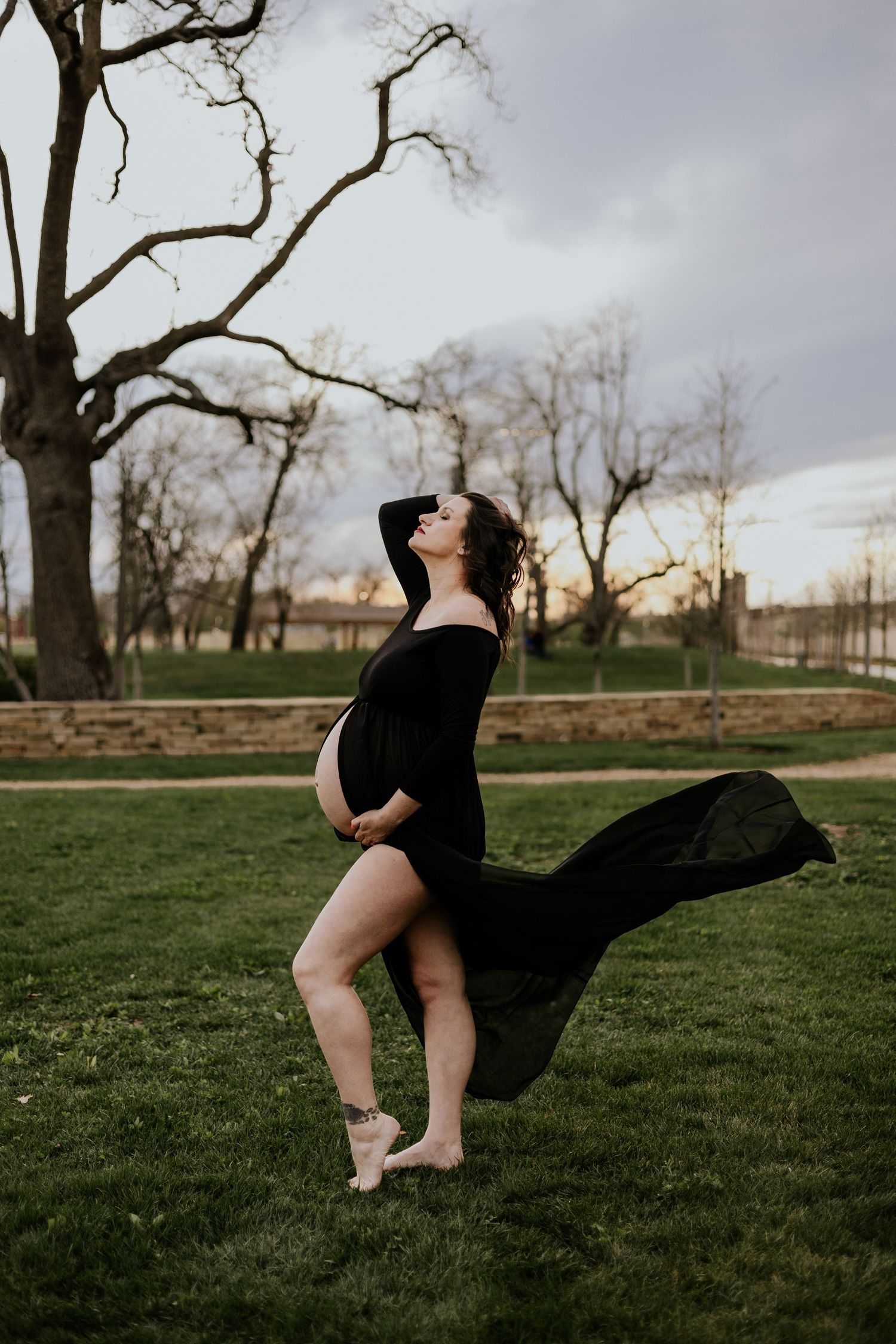 Pregnant woman in black flowing maternity gown poses outdoors in park with fabric billowing in wind.